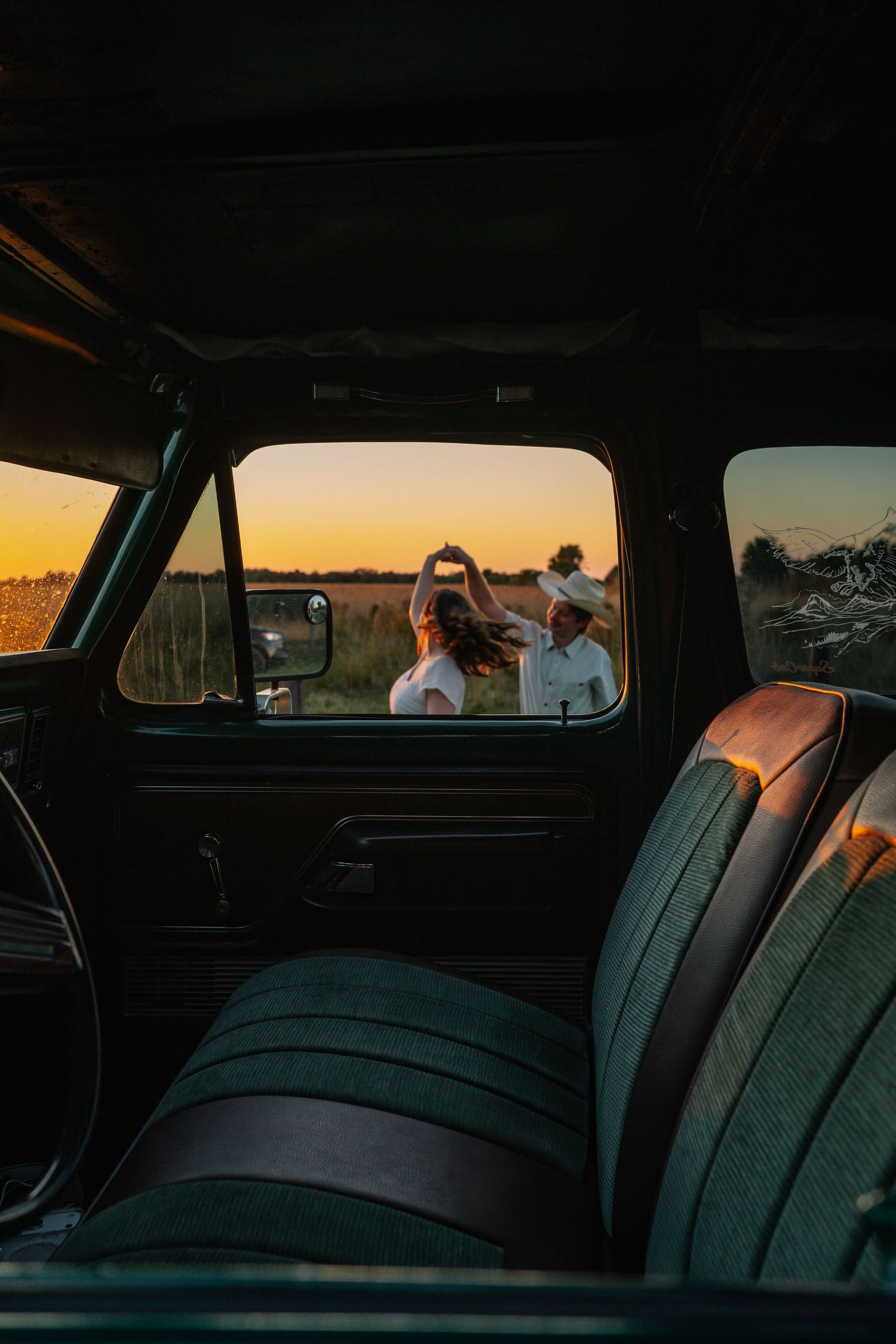 Romantic couple silhouetted at sunset viewed through the window of a vintage truck cab in an open field.