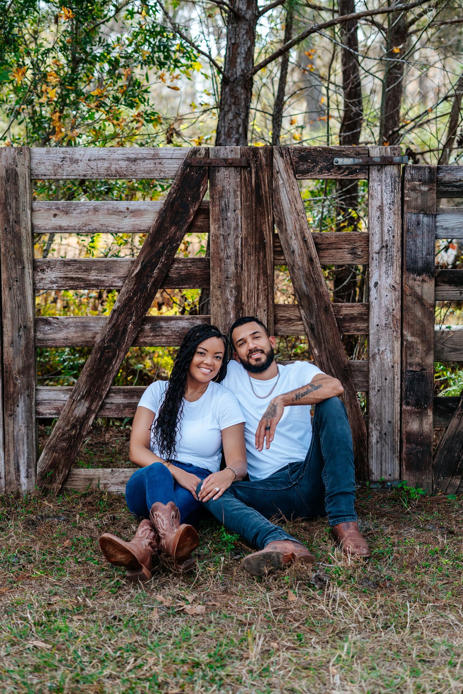 Smiling couple in matching white tees and jeans sitting together against a rustic wooden fence outdoors.