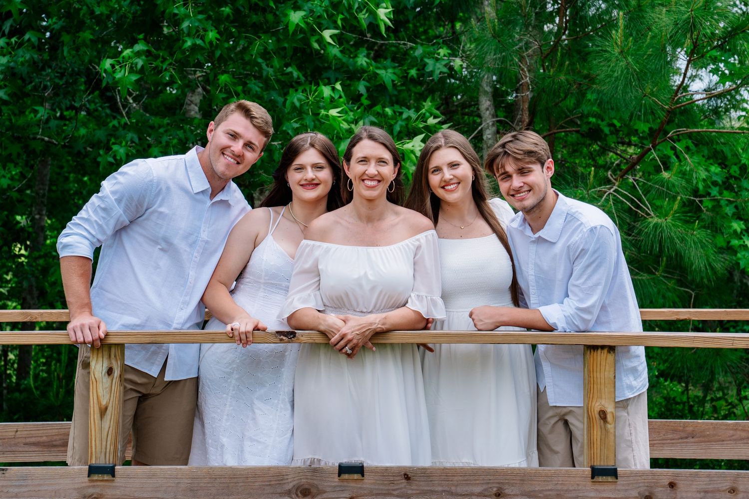 Five smiling people in white outfits pose together on a wooden bridge surrounded by lush green trees.
