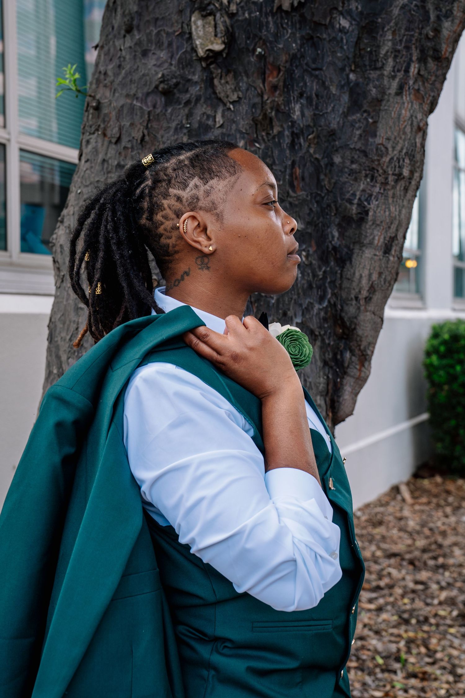 Person with dreadlocks in green suit vest and white shirt leaning against tree outdoors.