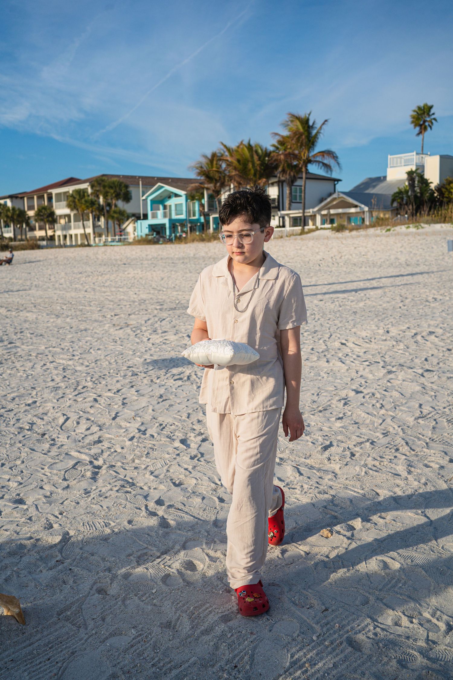 Person in beige outfit and red shoes walking on sandy beach with coastal houses and palm trees in background.