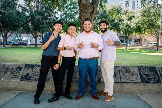 Group of people standing together in casual attire against an outdoor background with trees and buildings.