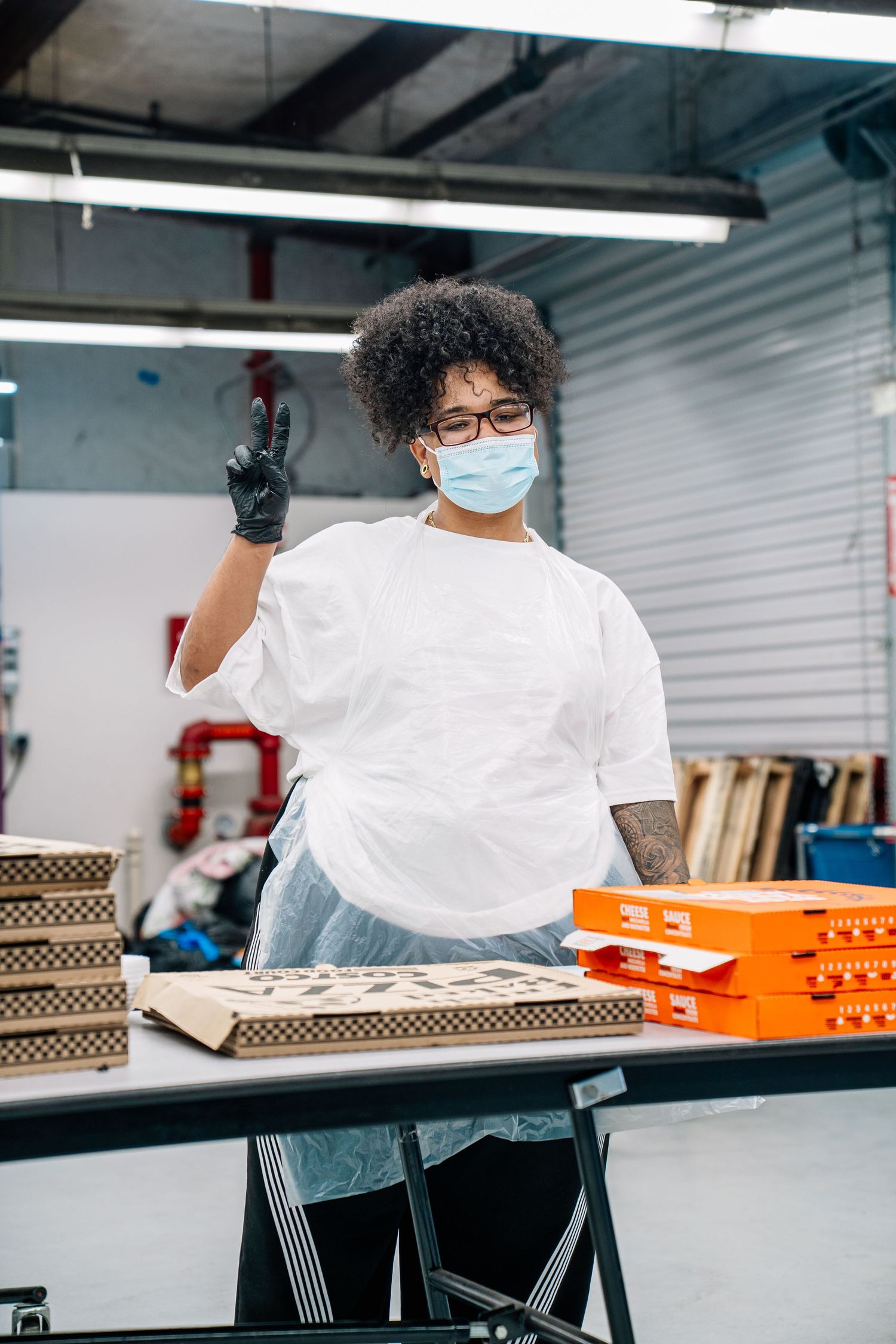 Worker in mask and gloves gives peace sign near cardboard boxes in a warehouse setting.