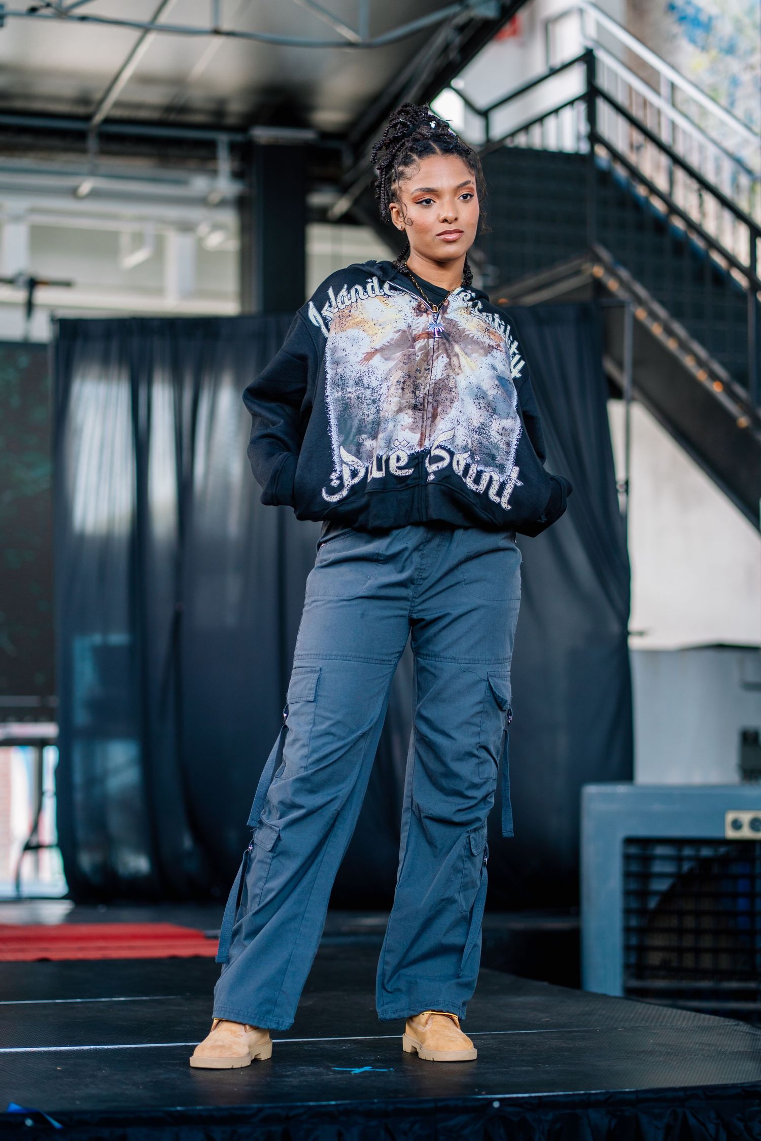 Model wearing graphic black sweatshirt and gray cargo pants on fashion runway.