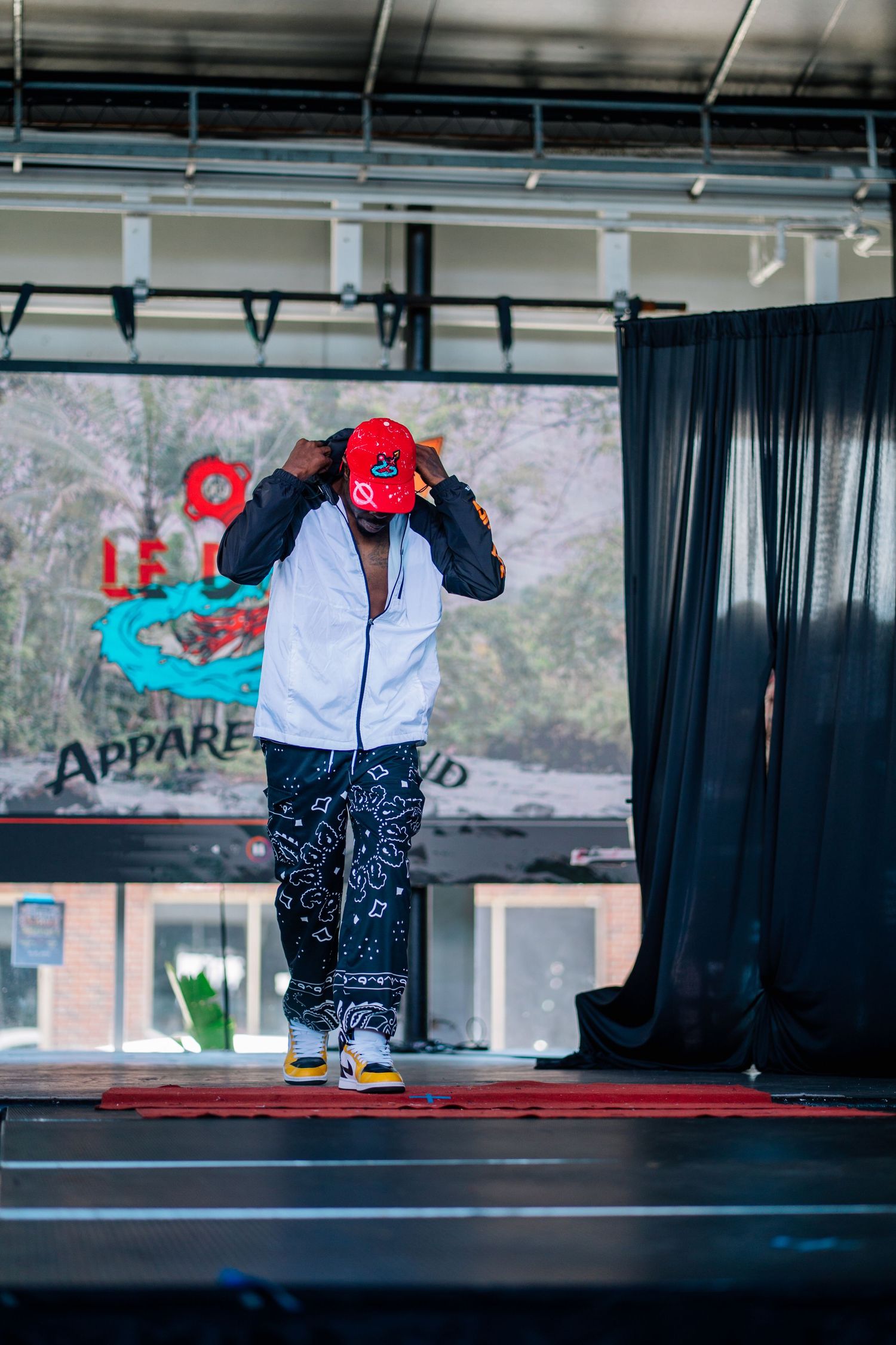 A performer on stage wearing a white hoodie, red cap, bandana print pants, and yellow sneakers at an outdoor apparel event.
