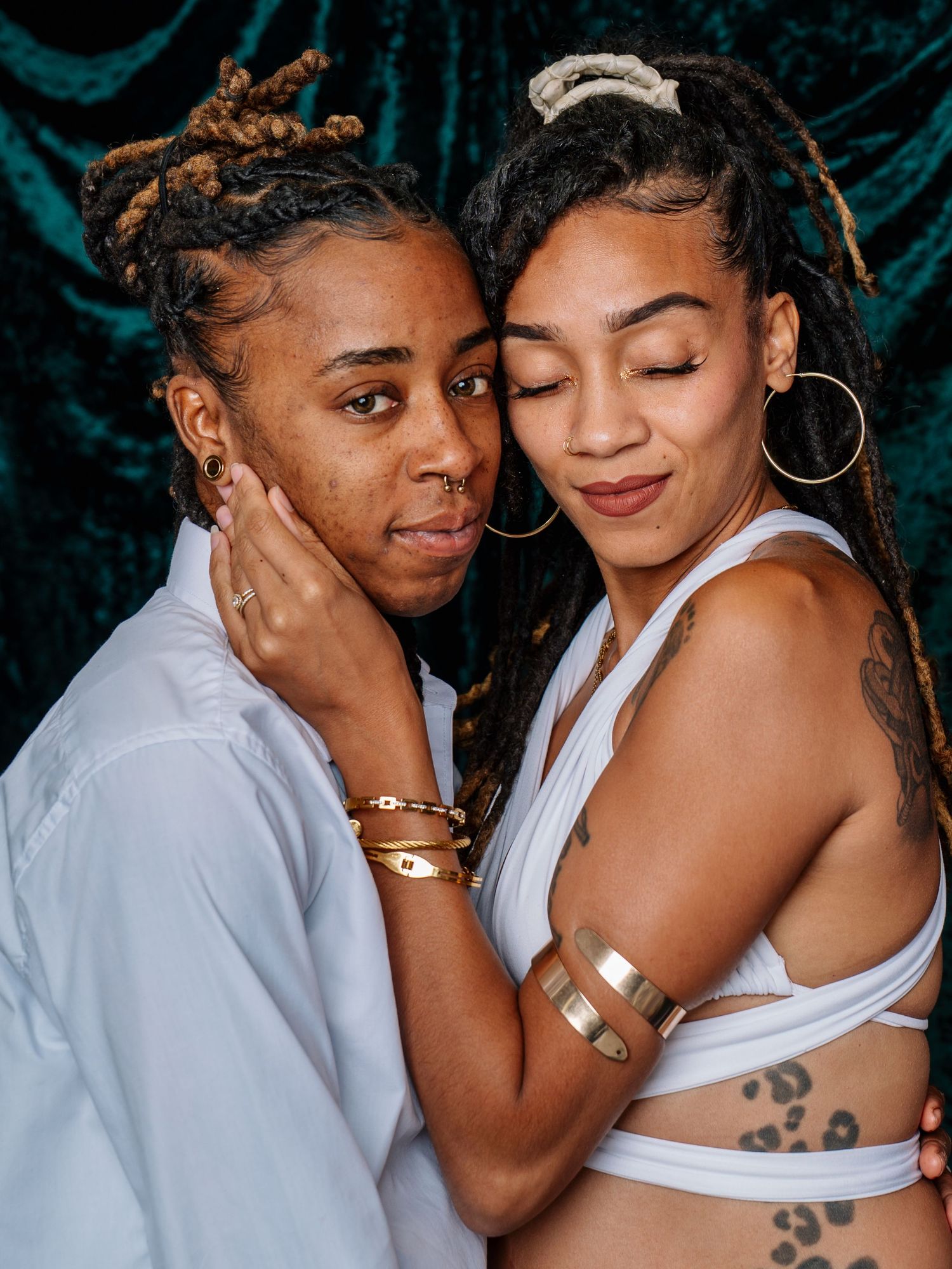 Two women with dreadlocks wearing white outfits and gold jewelry pose closely together against a teal velvet backdrop.