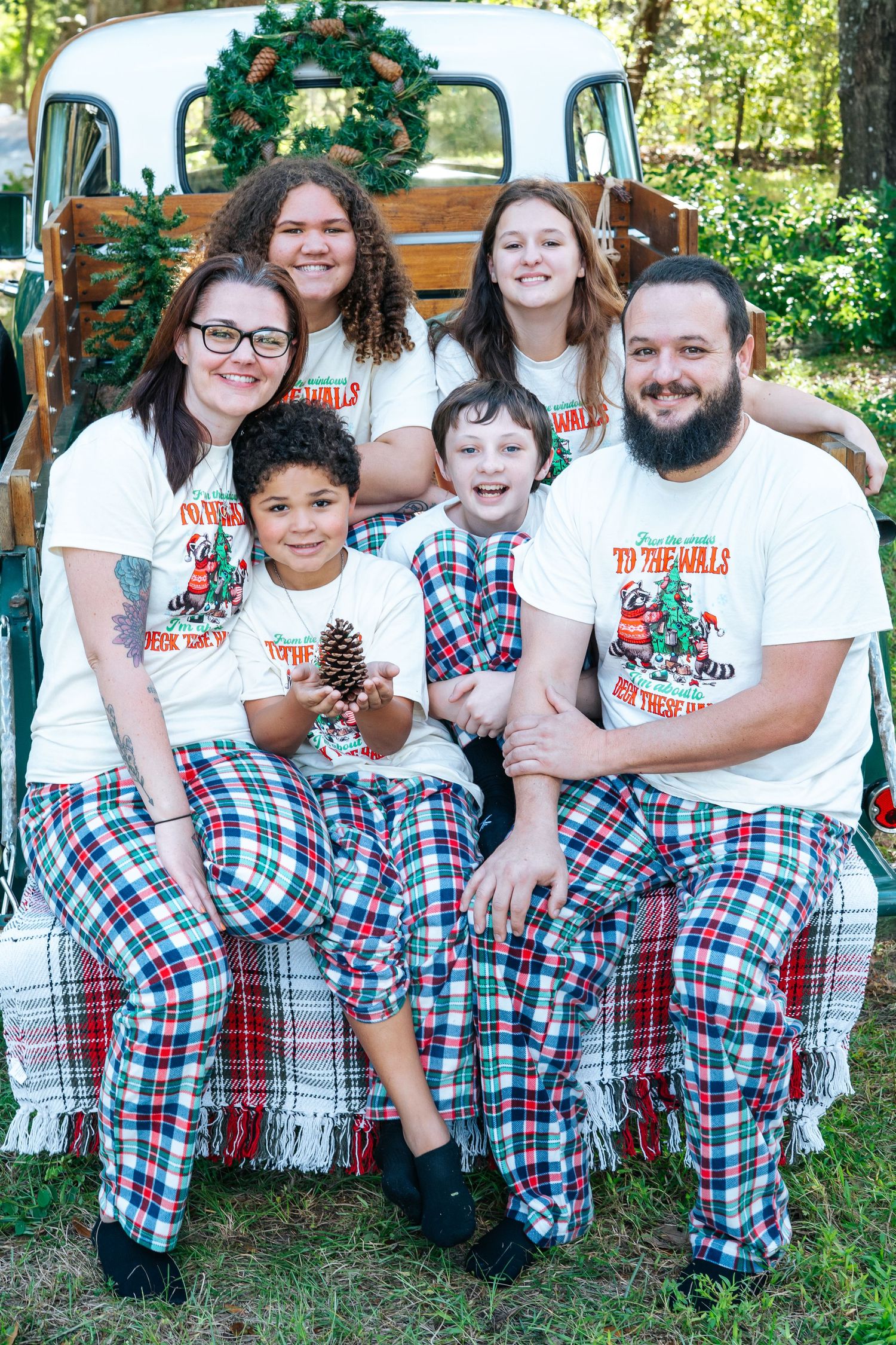 Family in matching Christmas t-shirts and plaid pants poses with holiday decorations.