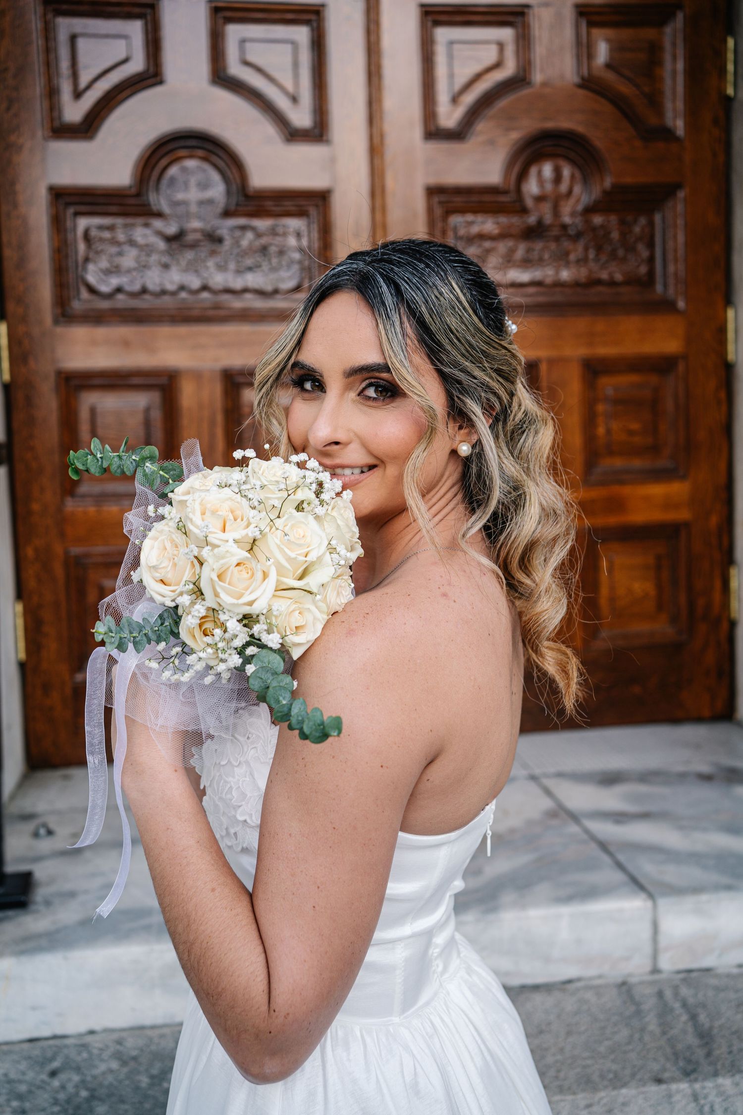 Bride in strapless white gown holds white rose bouquet, smiling over shoulder before ornate wooden church doors.