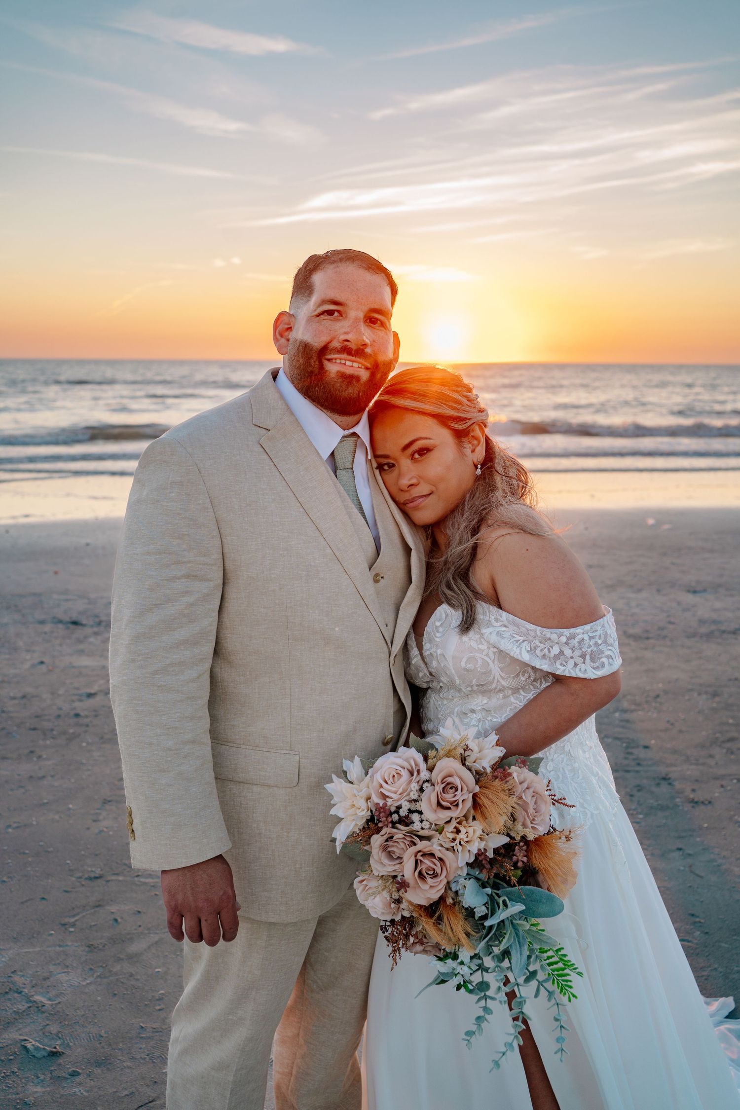 Bride and groom pose on beach at sunset, she holds bouquet, he wears tan suit, ocean waves in background.