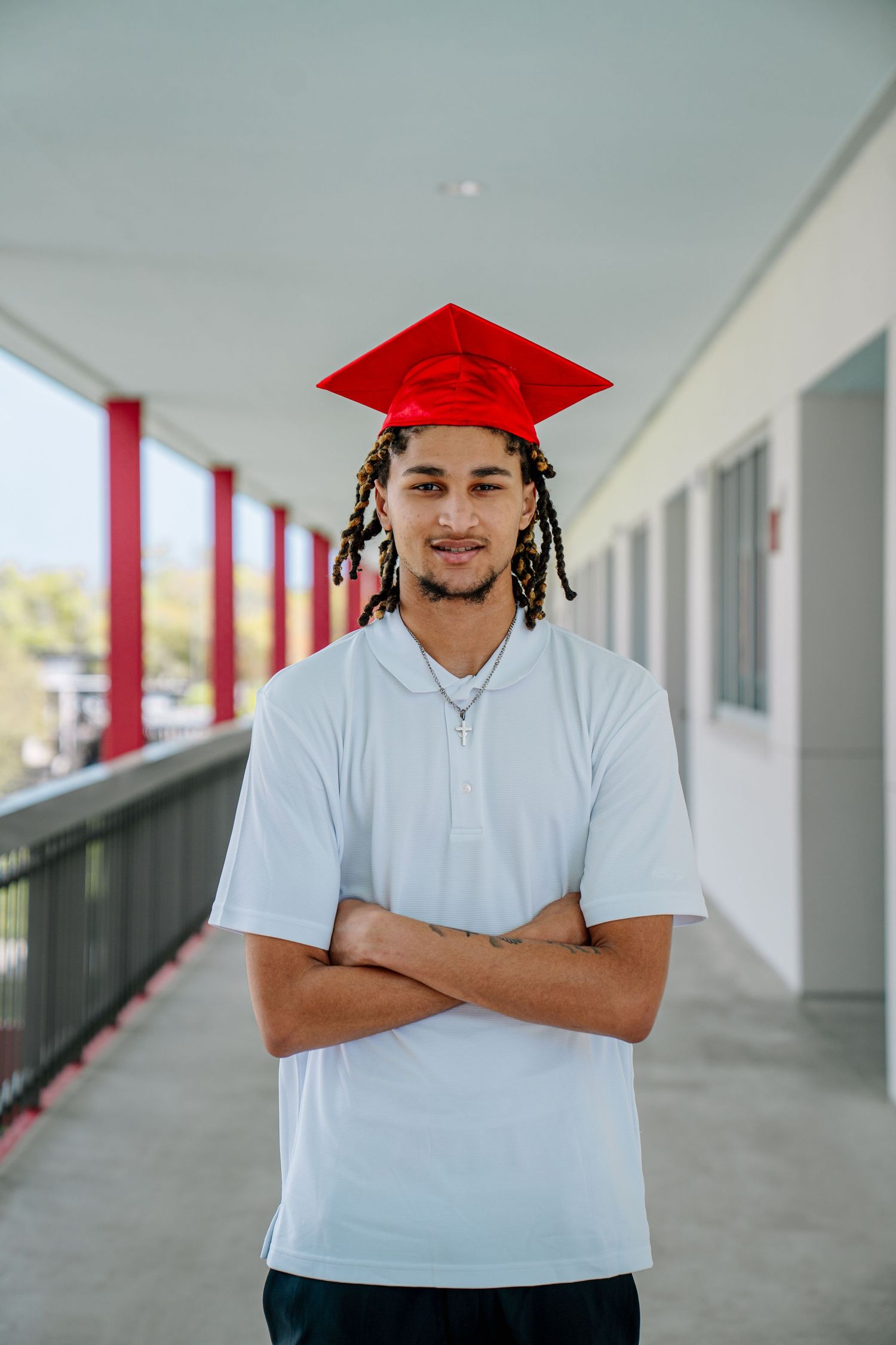 Young man wearing a red graduation cap and white polo shirt stands with arms crossed on a school corridor.
