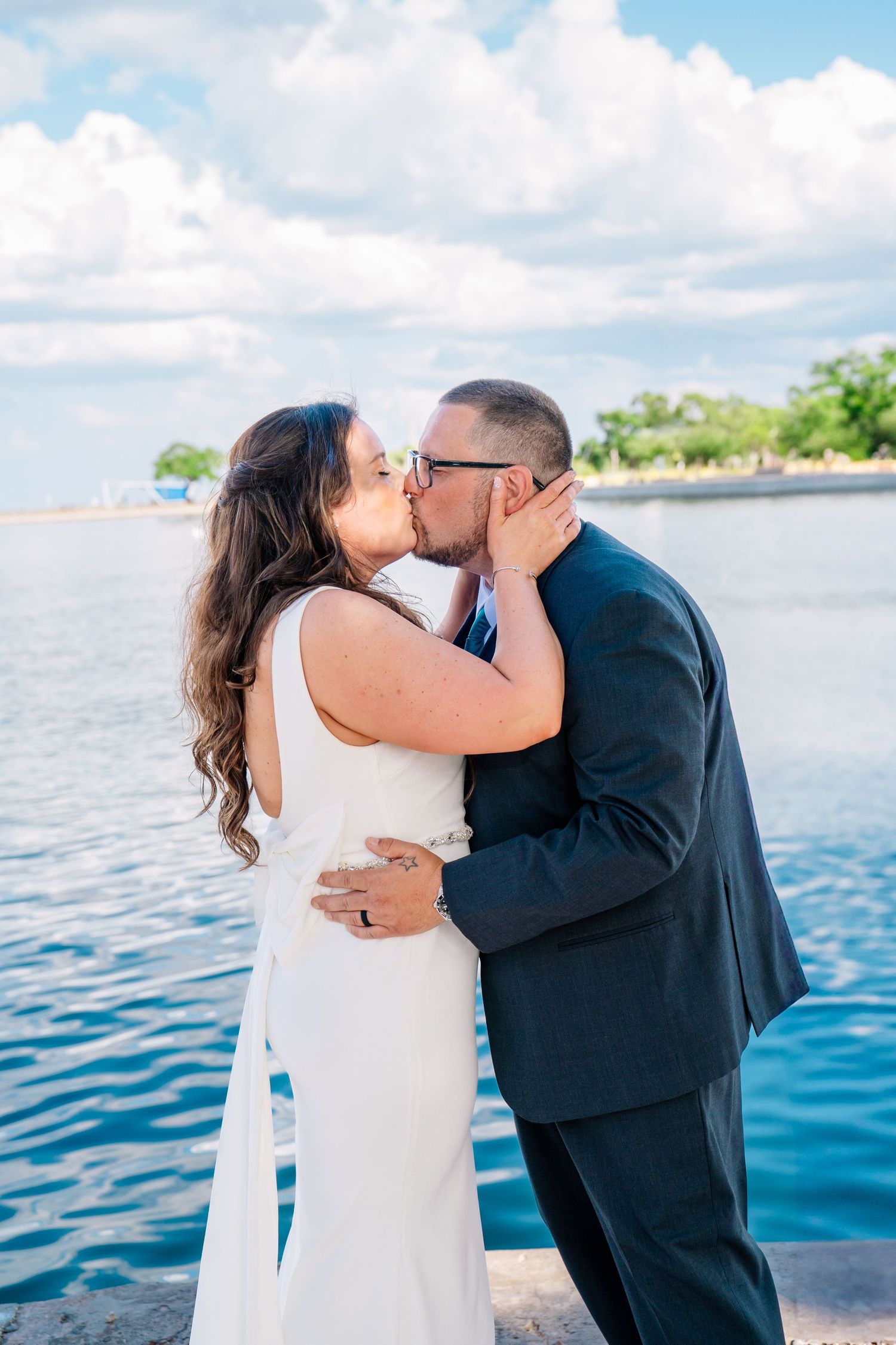Bride and groom share a romantic kiss by the waterfront, dressed in white gown and dark suit on their wedding day.