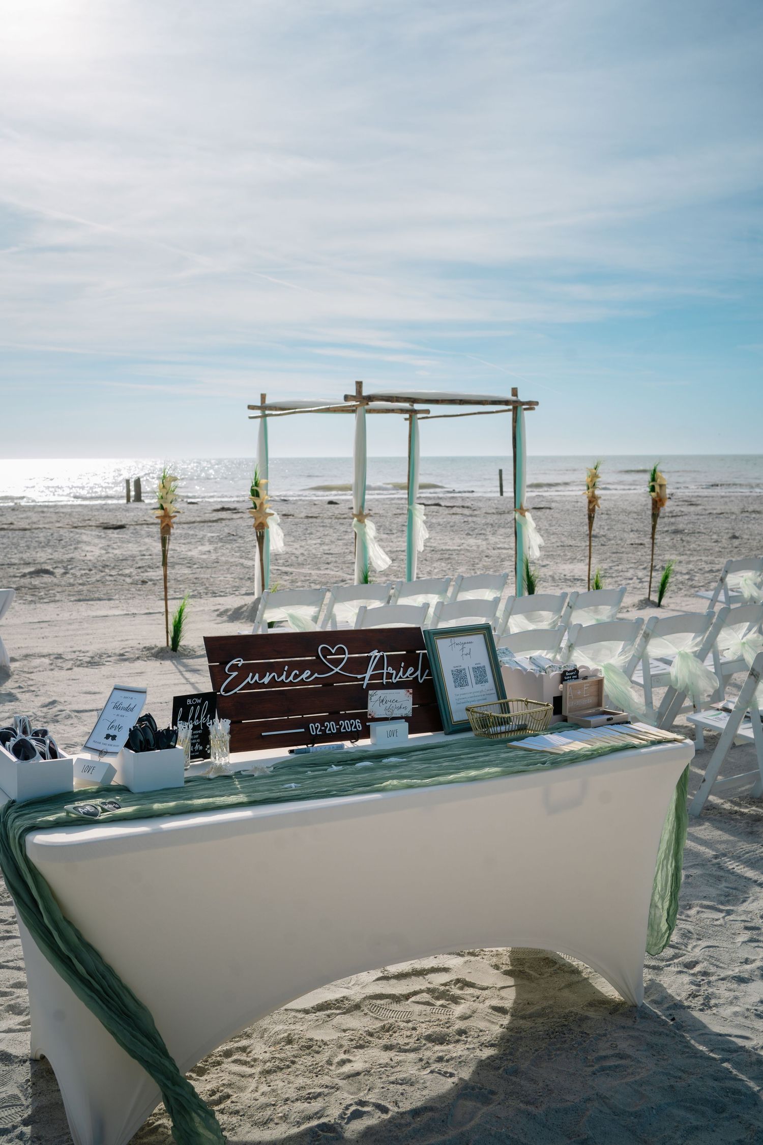 A beach wedding setup with tiki torches and white chairs arranged in rows on sandy shores under a blue sky.