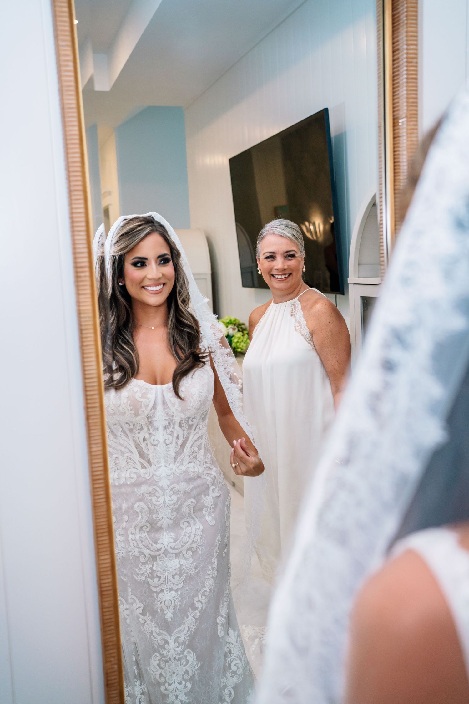 Bride in lace wedding gown and veil smiles with bridesmaid in white dress, reflected in gold-framed mirror.