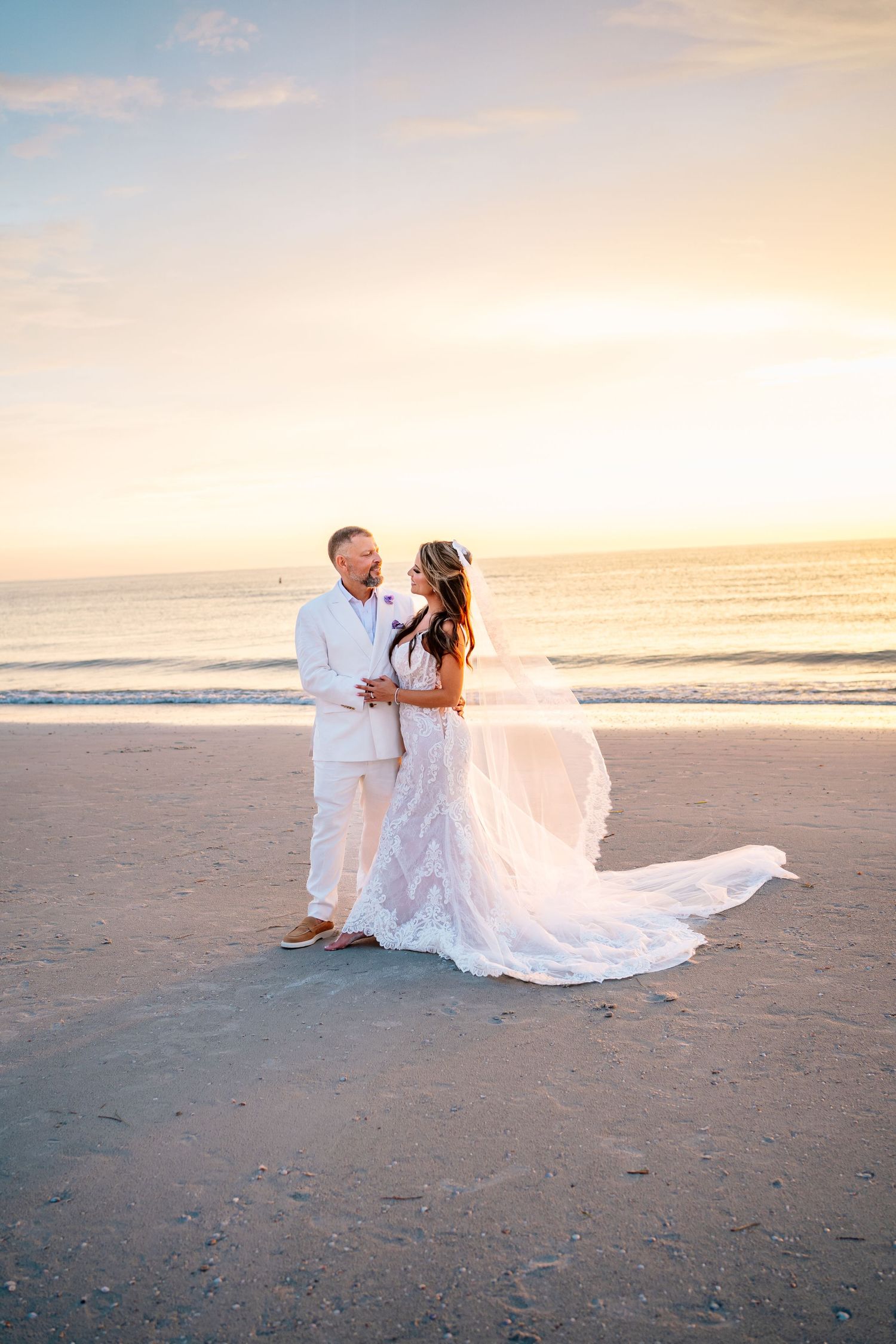 Bride and groom embrace on a sandy beach at sunset, her white lace gown and veil flowing in the breeze.