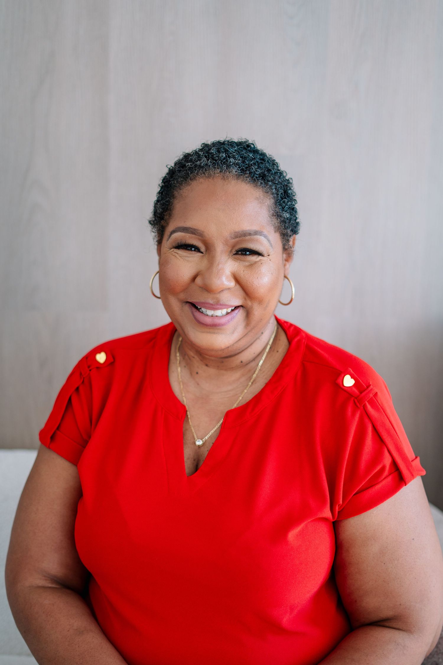 Smiling middle-aged Black woman with short gray hair wearing a red blouse and hoop earrings against a neutral background.