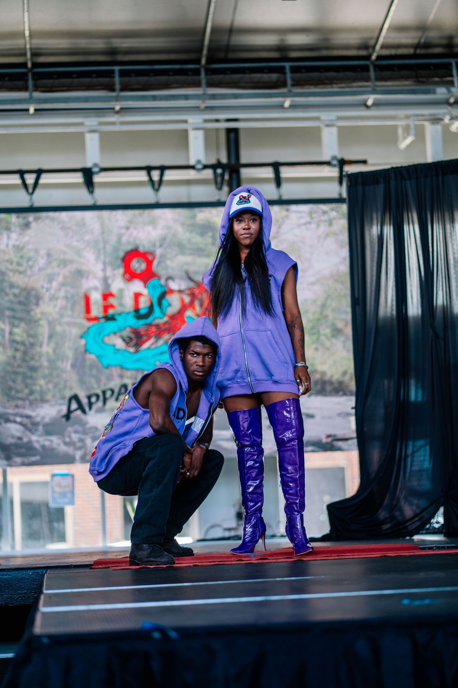 Two models on a runway wearing matching purple outfits with thigh-high boots at a fashion show.