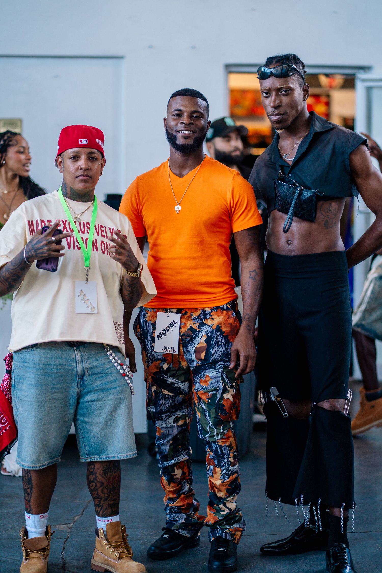 Three stylish men pose together at an event, wearing bold streetwear including orange tee, camo pants, and edgy black outfit.