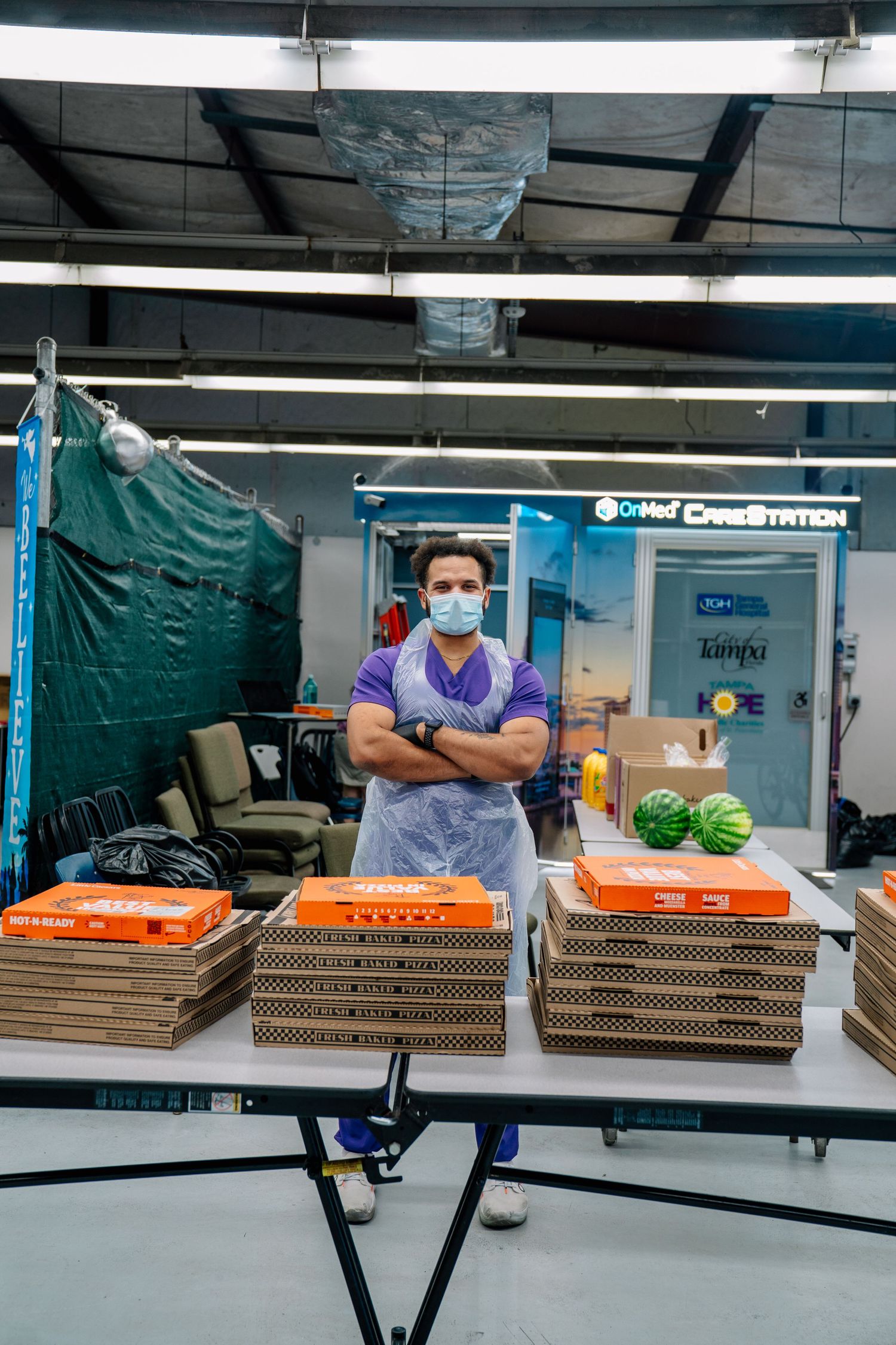 Man in mask and apron stands arms crossed behind table with pizza boxes and watermelons at a food distribution center.