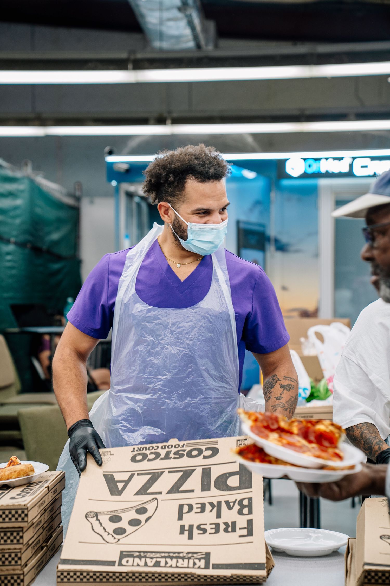 A masked worker in a purple shirt and apron carries Costco Kirkland pizza boxes at a food service event.