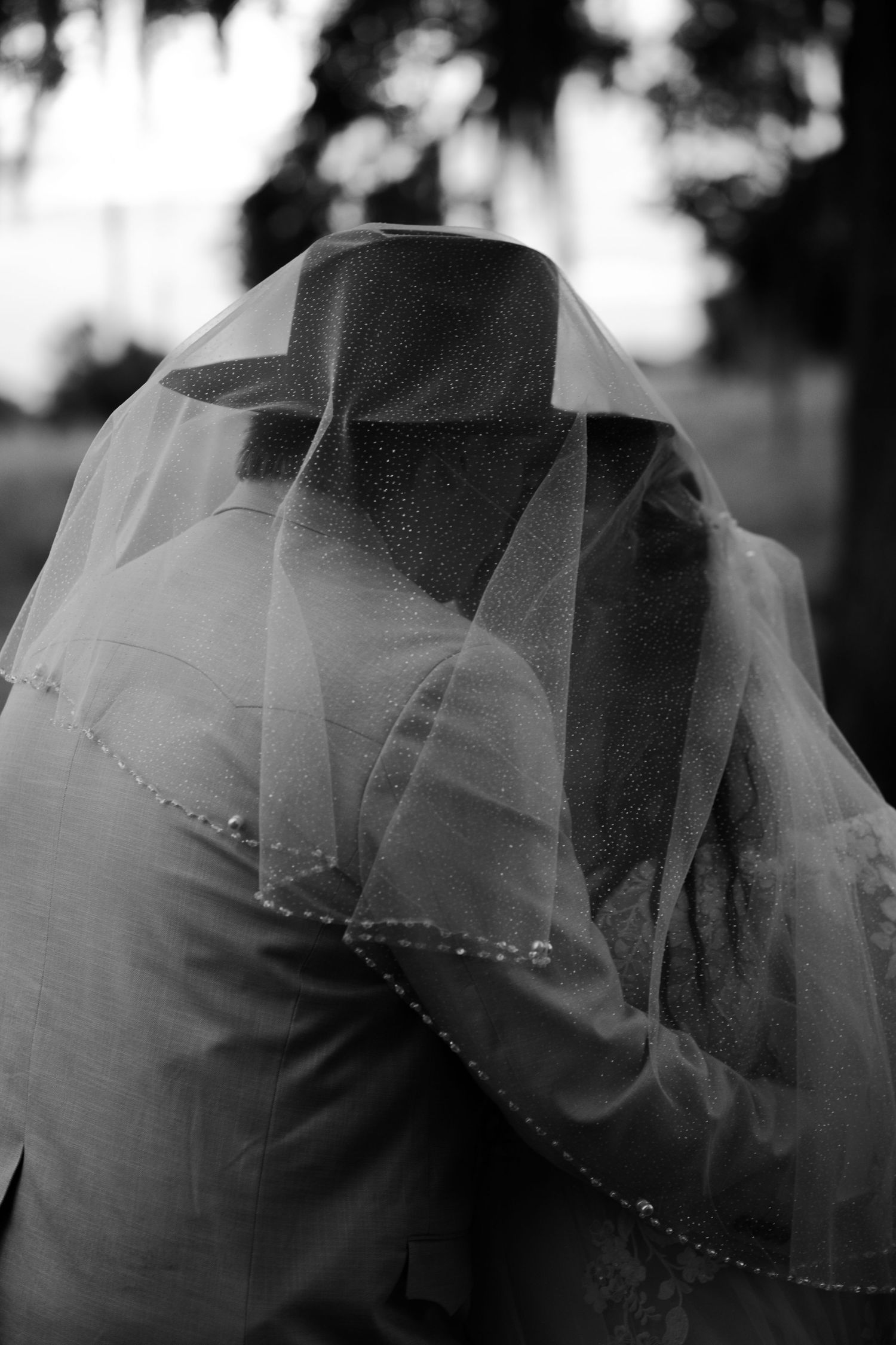 A black and white wedding photograph showing a delicate veil draped over a suit-clad figure in soft focus.