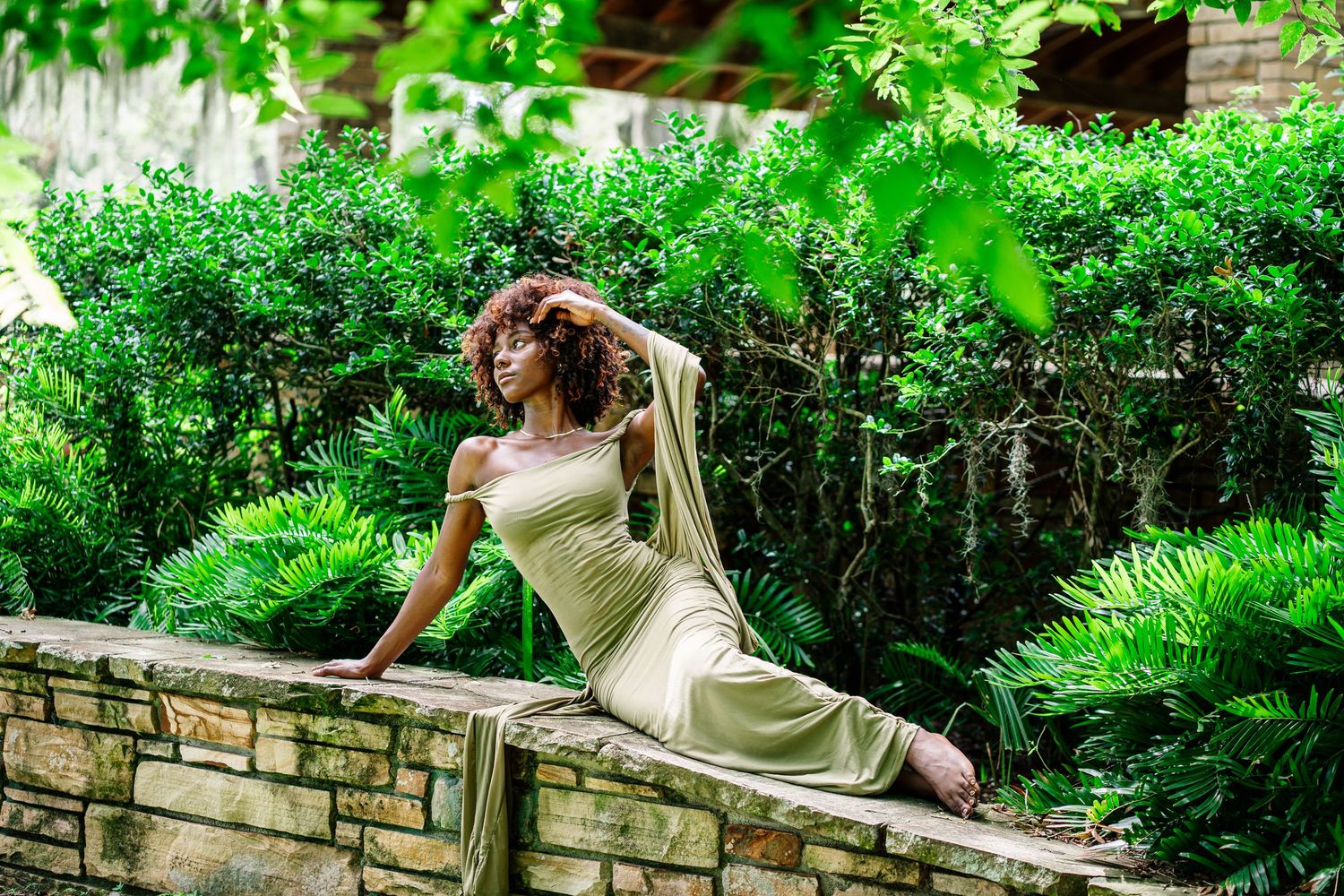 Woman in elegant green dress poses gracefully on stone wall surrounded by lush garden greenery.
