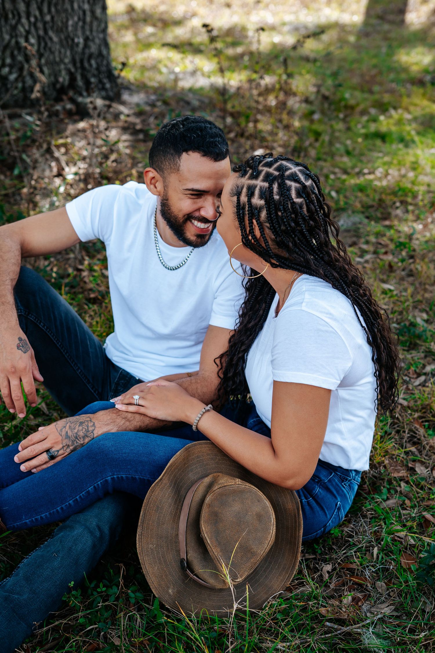 Happy couple sitting on grass laughing together with a brown hat nearby in a sunlit forest.