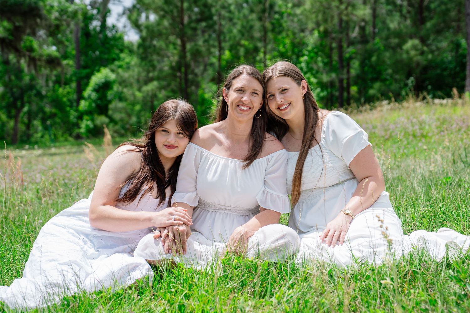 Three women in white dresses sitting together on green grass in a sunny outdoor park setting.