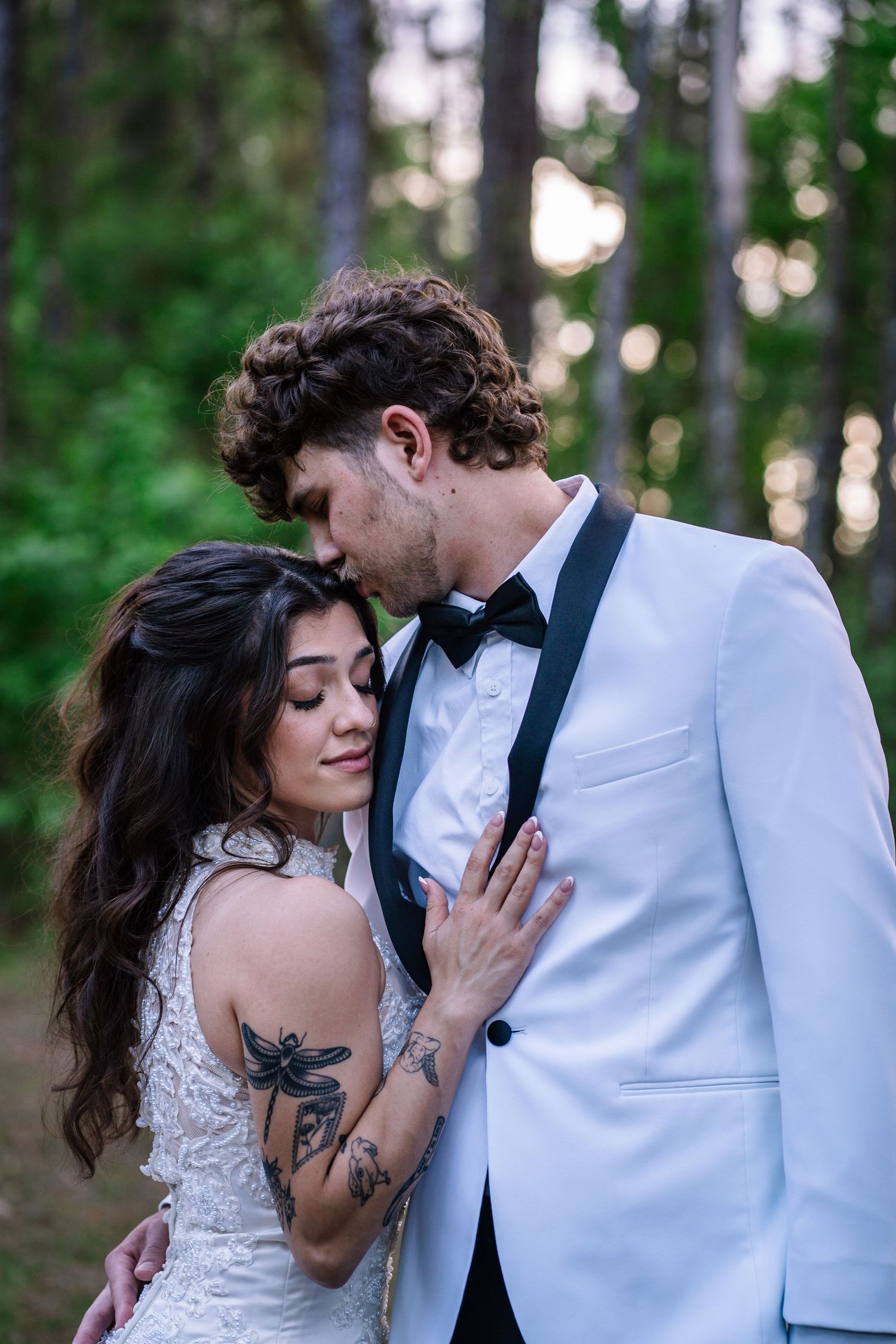 Bride and groom embrace in forest, she wears lace dress with tattoos, he wears white tuxedo with bow tie.