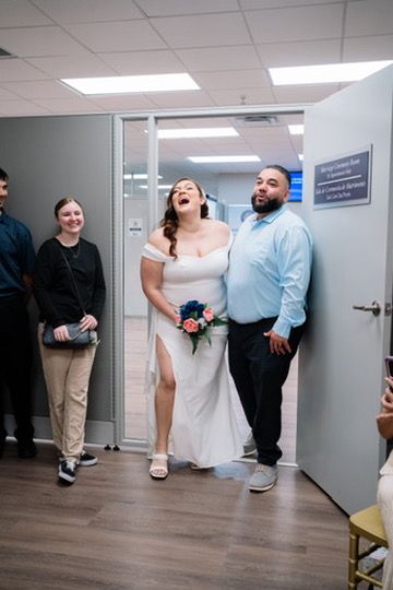 A joyful wedding moment captured in an office hallway as a bride in white laughs while holding a small bouquet.