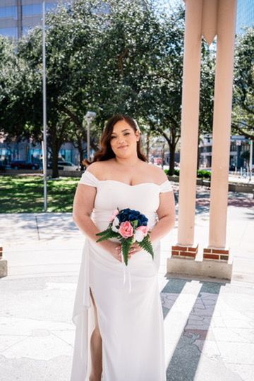 Bride in elegant white off-shoulder gown poses with pink and blue bouquet under covered outdoor walkway.