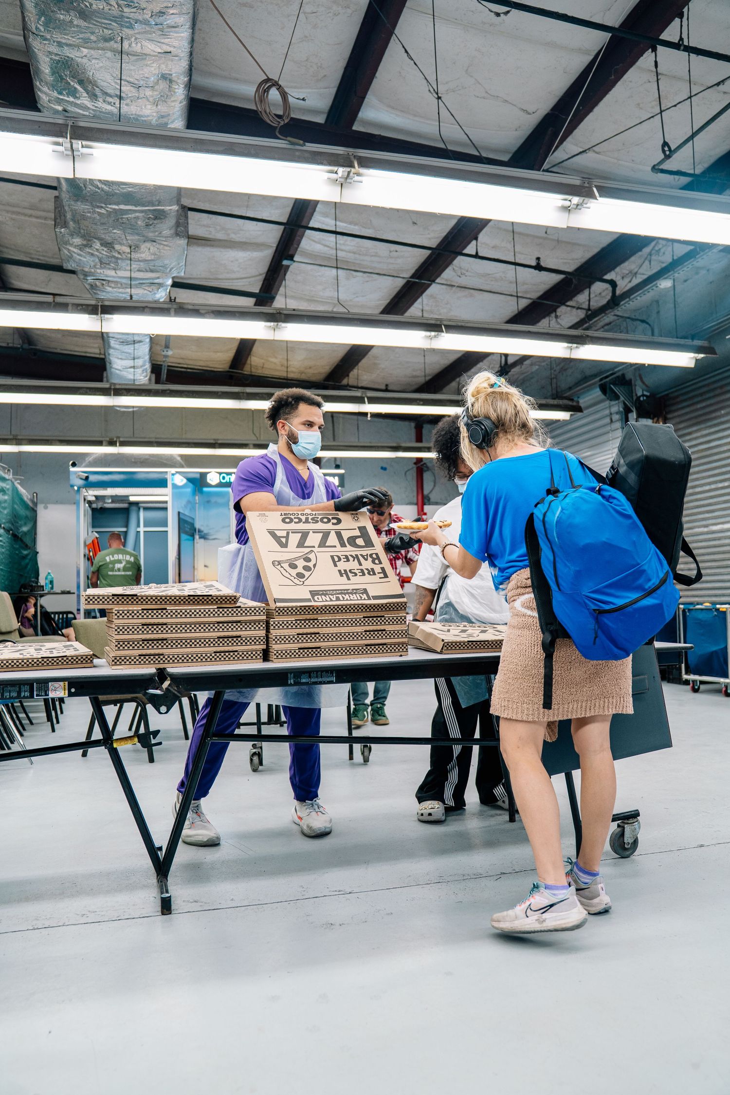 Masked volunteer hands a pizza box to a woman with a backpack at an indoor food distribution center.