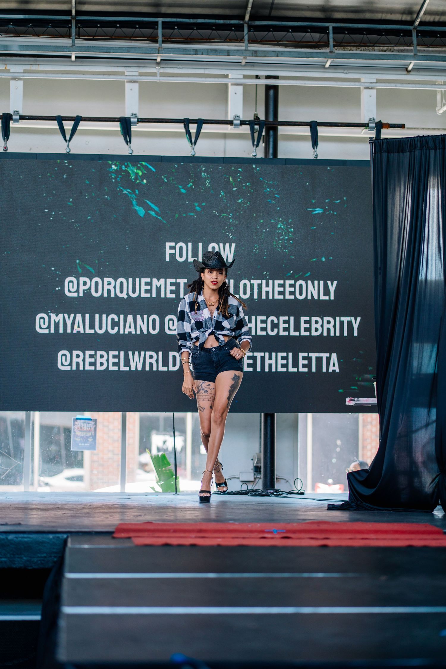 A model walks the runway at a fashion show with social media handles displayed on a large screen behind her.