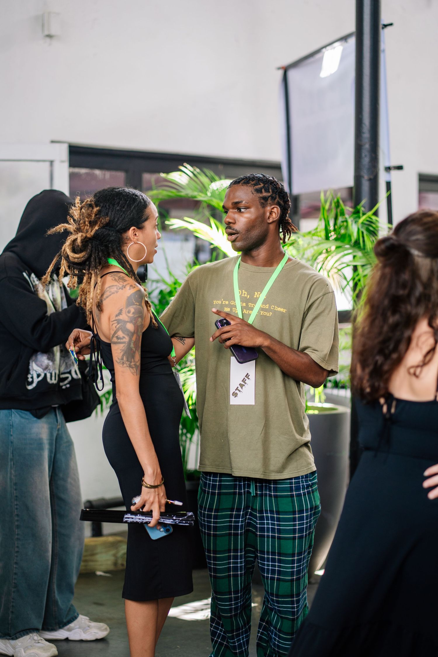 Staff member in plaid pants and green tee chats with tattooed woman at an indoor event with tropical plants.