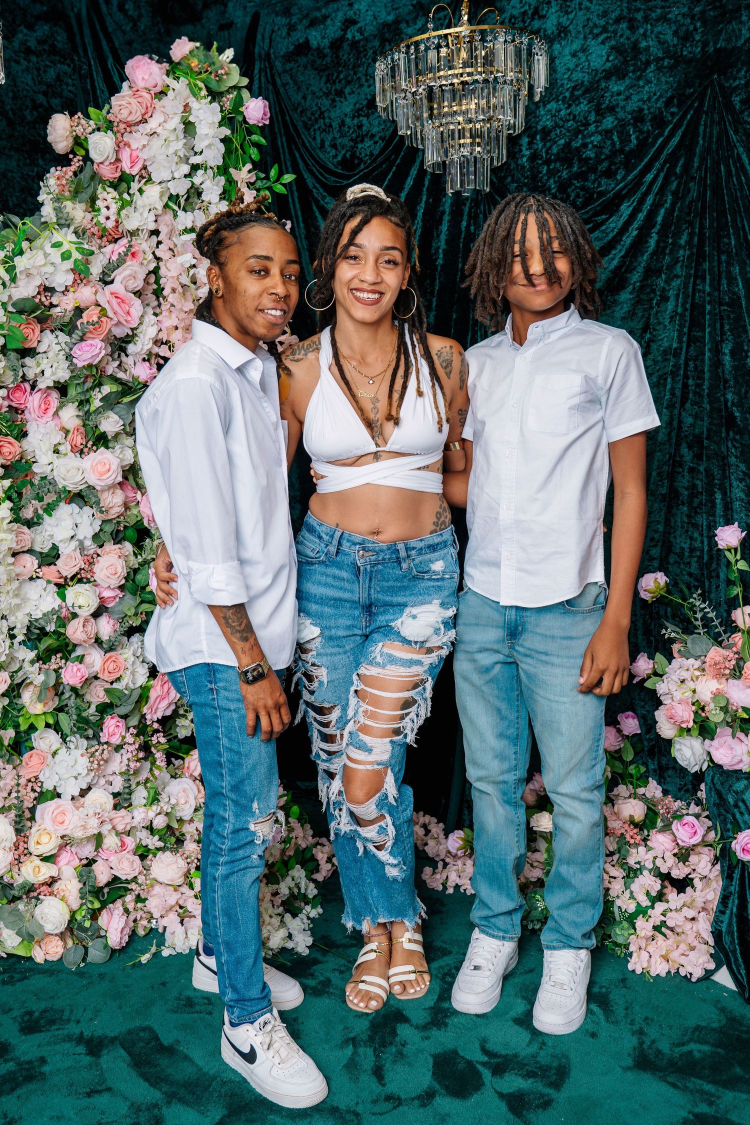 Three young people posing together in front of a floral backdrop with a chandelier, wearing white tops and jeans.