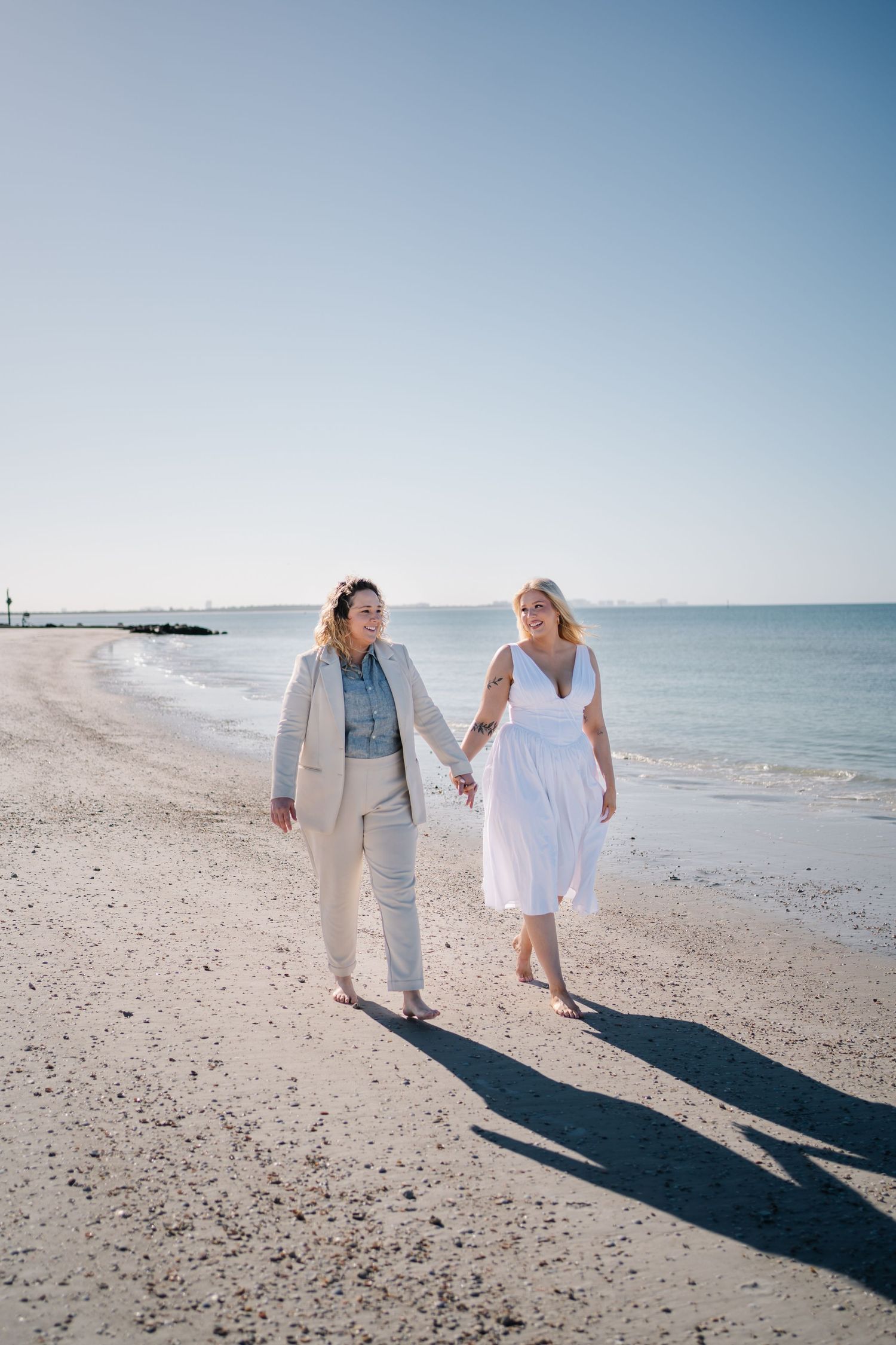 Two women holding hands walking barefoot on sunny beach, one in white dress and one in beige suit.