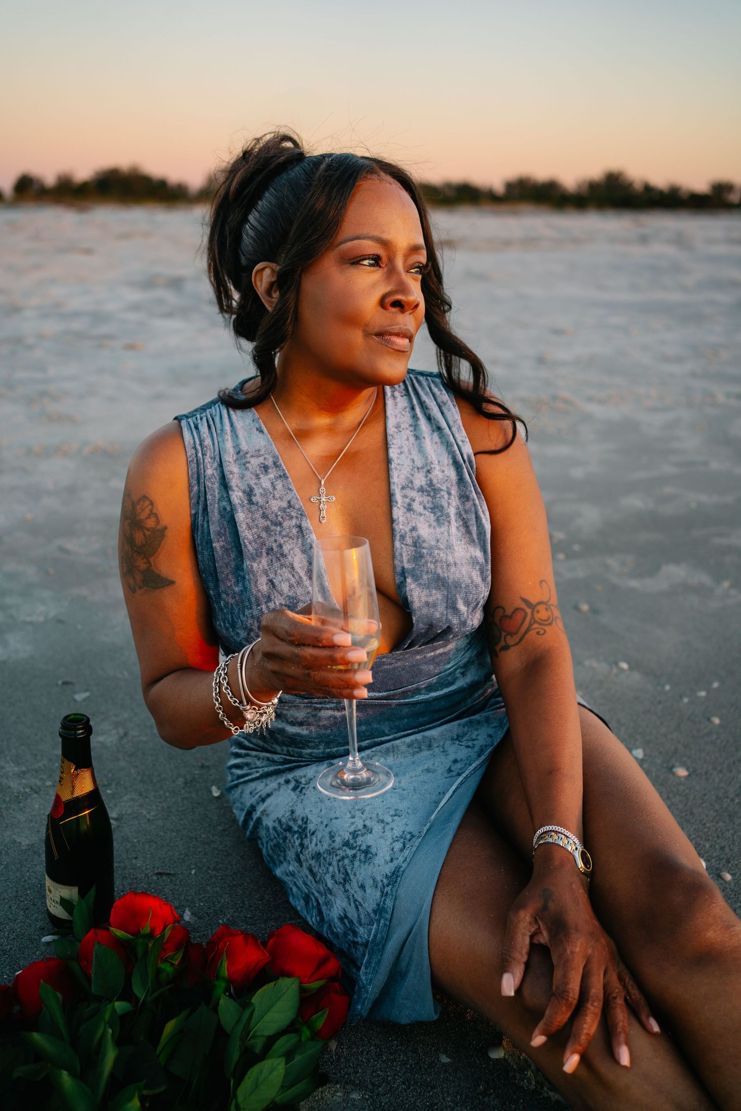 Woman in blue velvet dress holding champagne glass at sunset beach with roses and wine bottle.