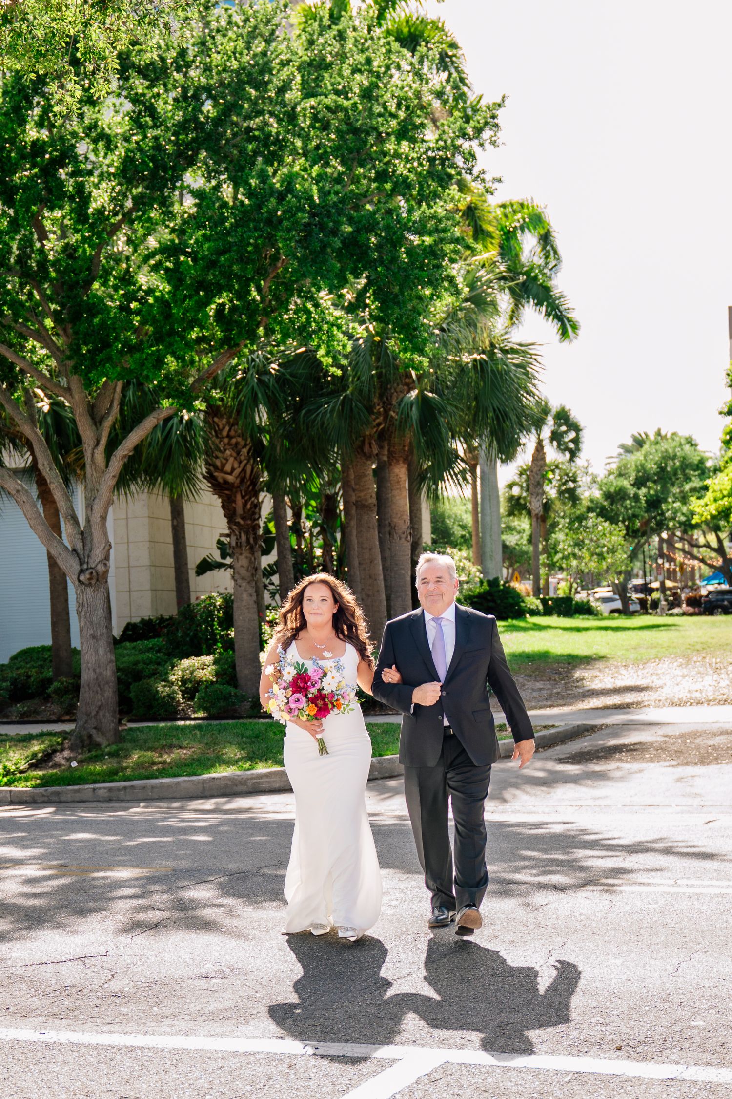 Bride in white gown holding colorful bouquet walks with escort past palm trees on sunny wedding day.