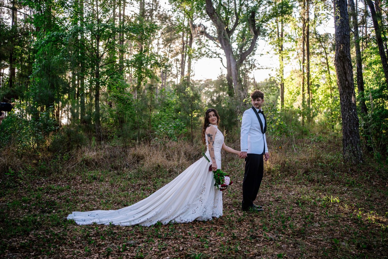 Bride in white gown and groom in white jacket hold hands in a sunlit forest clearing for wedding portraits.