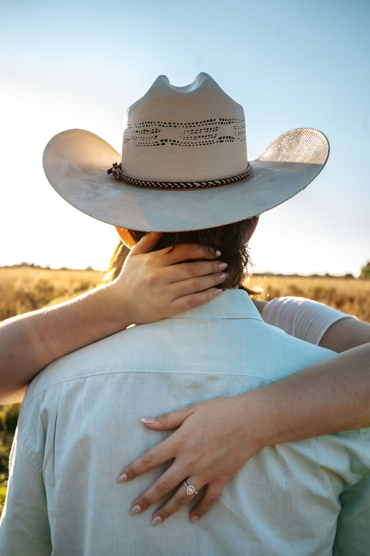Couple embracing in a golden field at sunset, man wearing a white cowboy hat and light blue shirt.