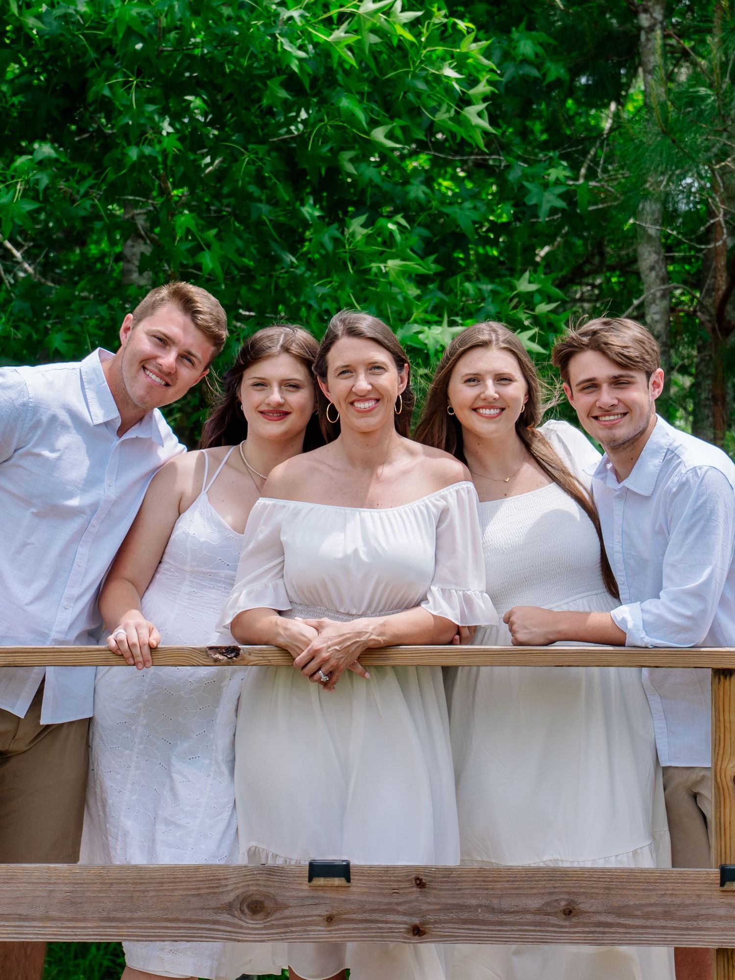 Five young adults in white outfits smiling together on a wooden deck surrounded by lush green trees.