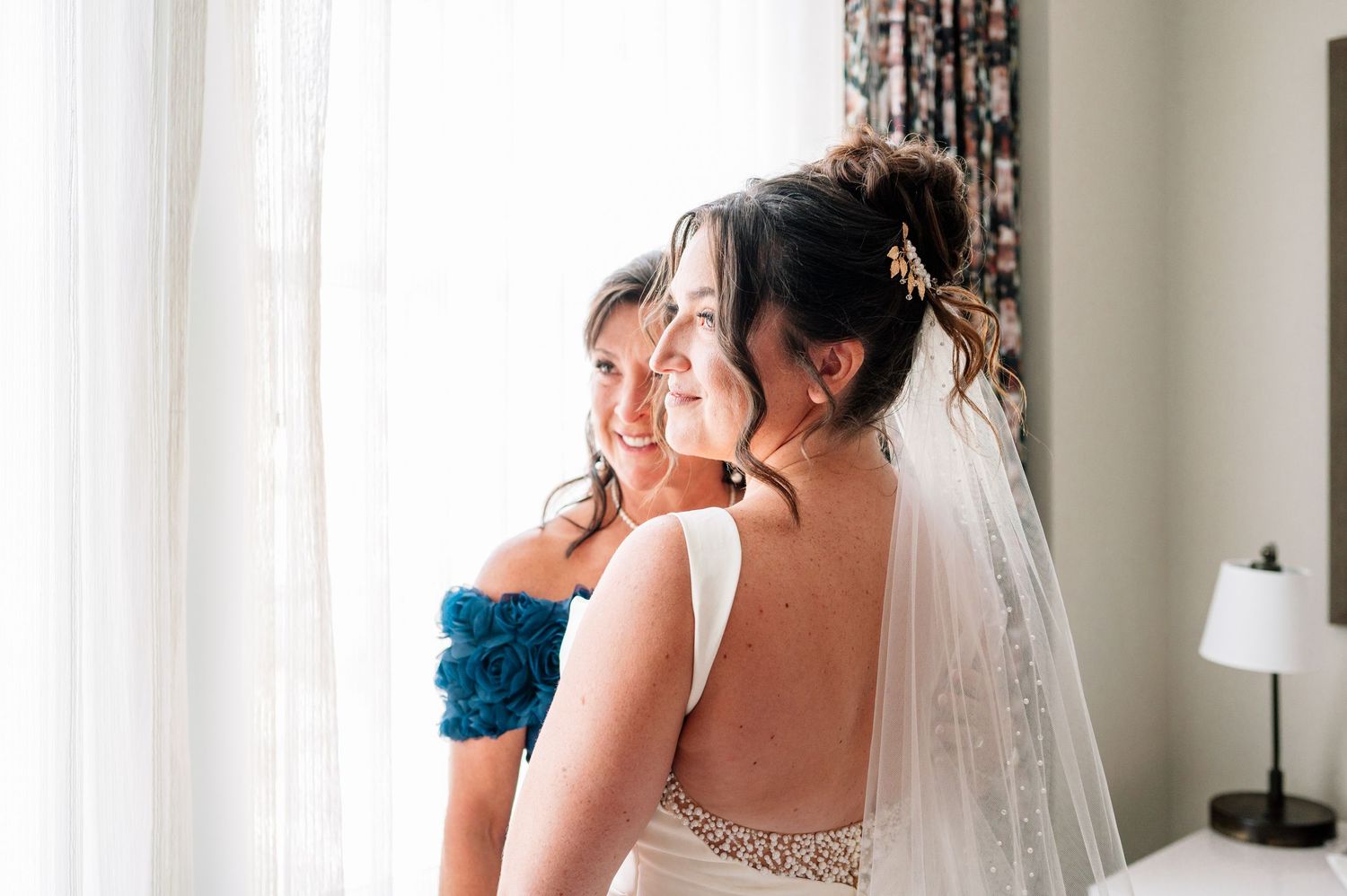 Bride in white gown and veil smiles with bridesmaid in teal dress by a bright window on wedding day.