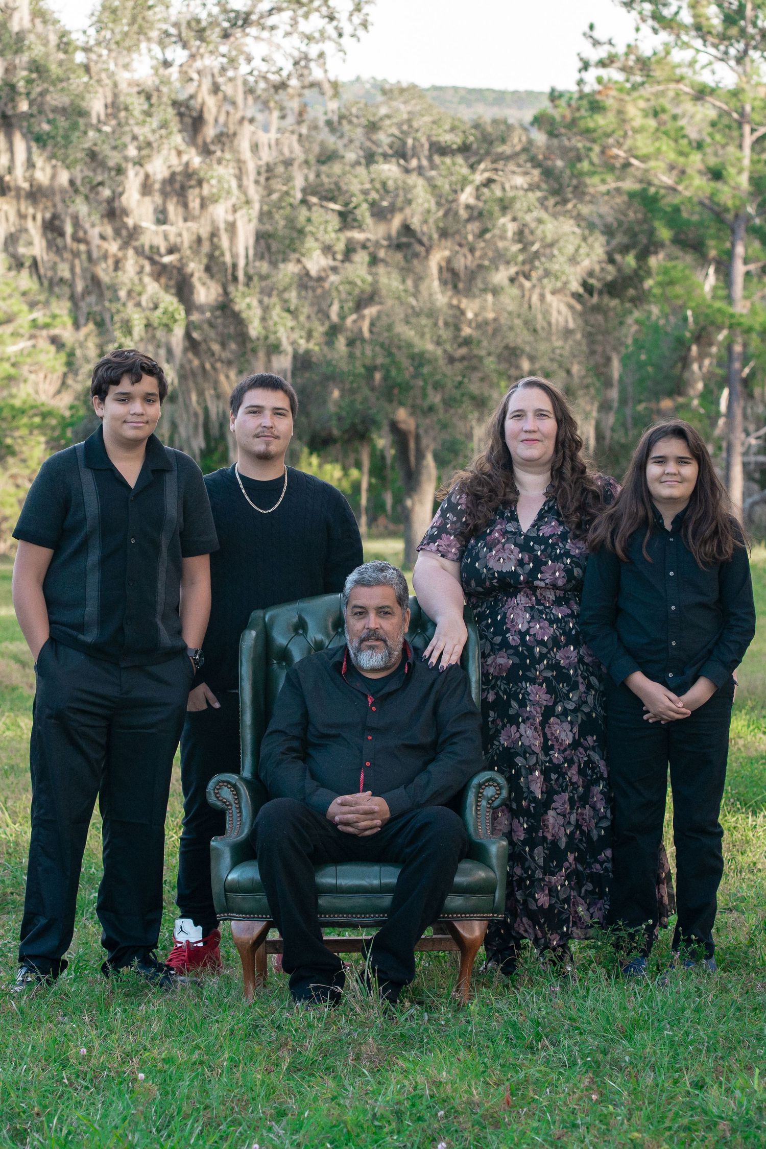 Family portrait in dark attire with vintage chair in a forest setting.