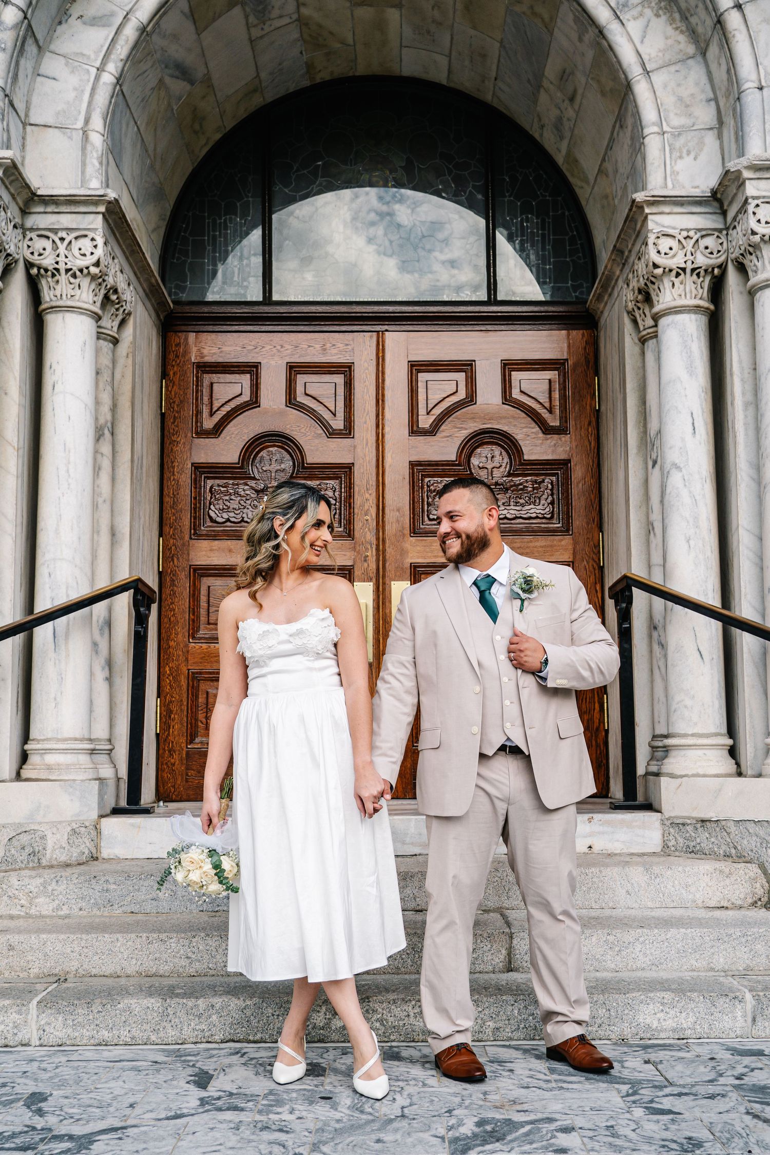 A couple in wedding attire shares playful moments in front of ornate stone archways and wooden doors.