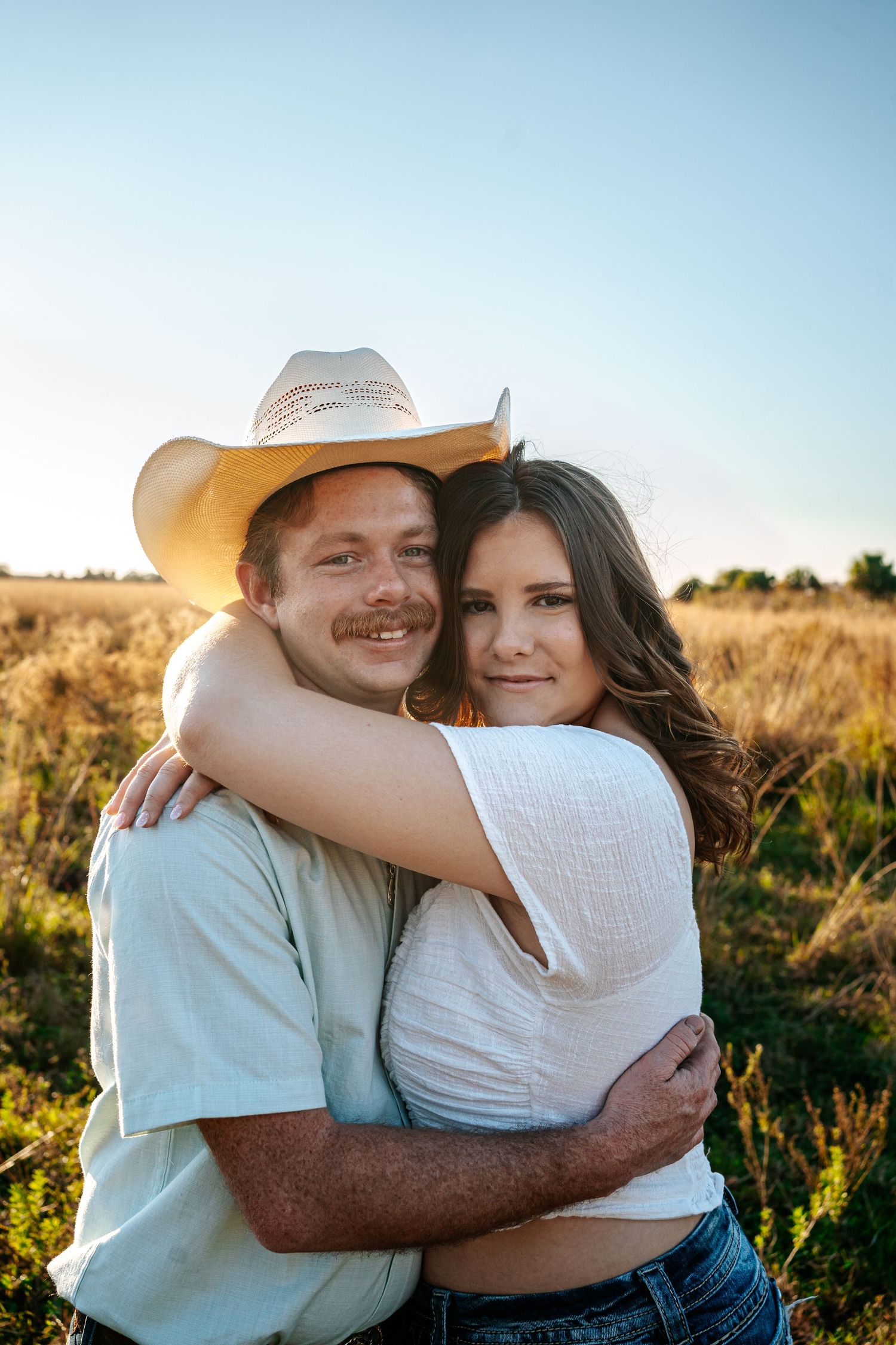 A couple embracing in a golden field at sunset, man wearing a cowboy hat and woman in a white top smiling warmly.