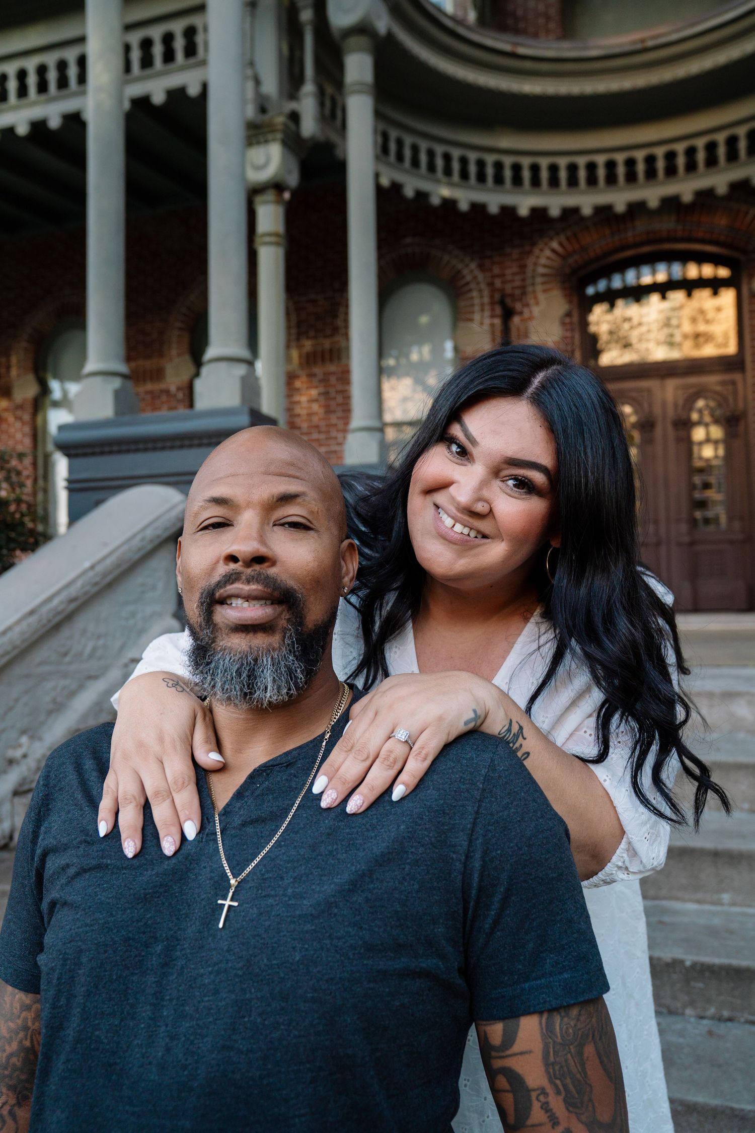 Smiling couple poses in front of a historic brick building, woman showing engagement ring and tattoos, man wearing cross necklace.