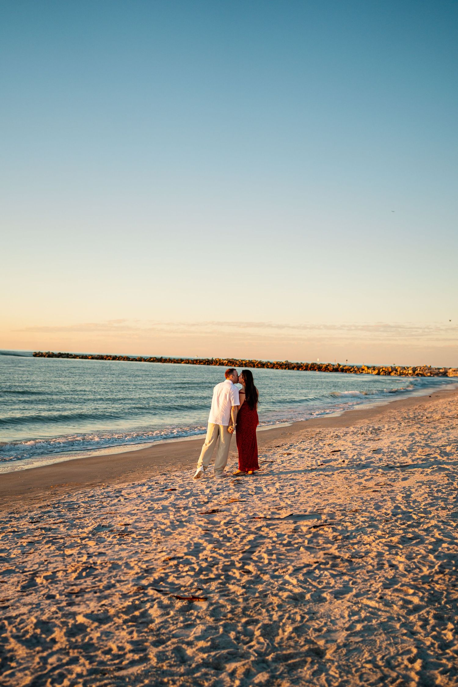 Couple embracing on a sandy beach at sunset, with calm ocean waves and a rocky pier in the background.