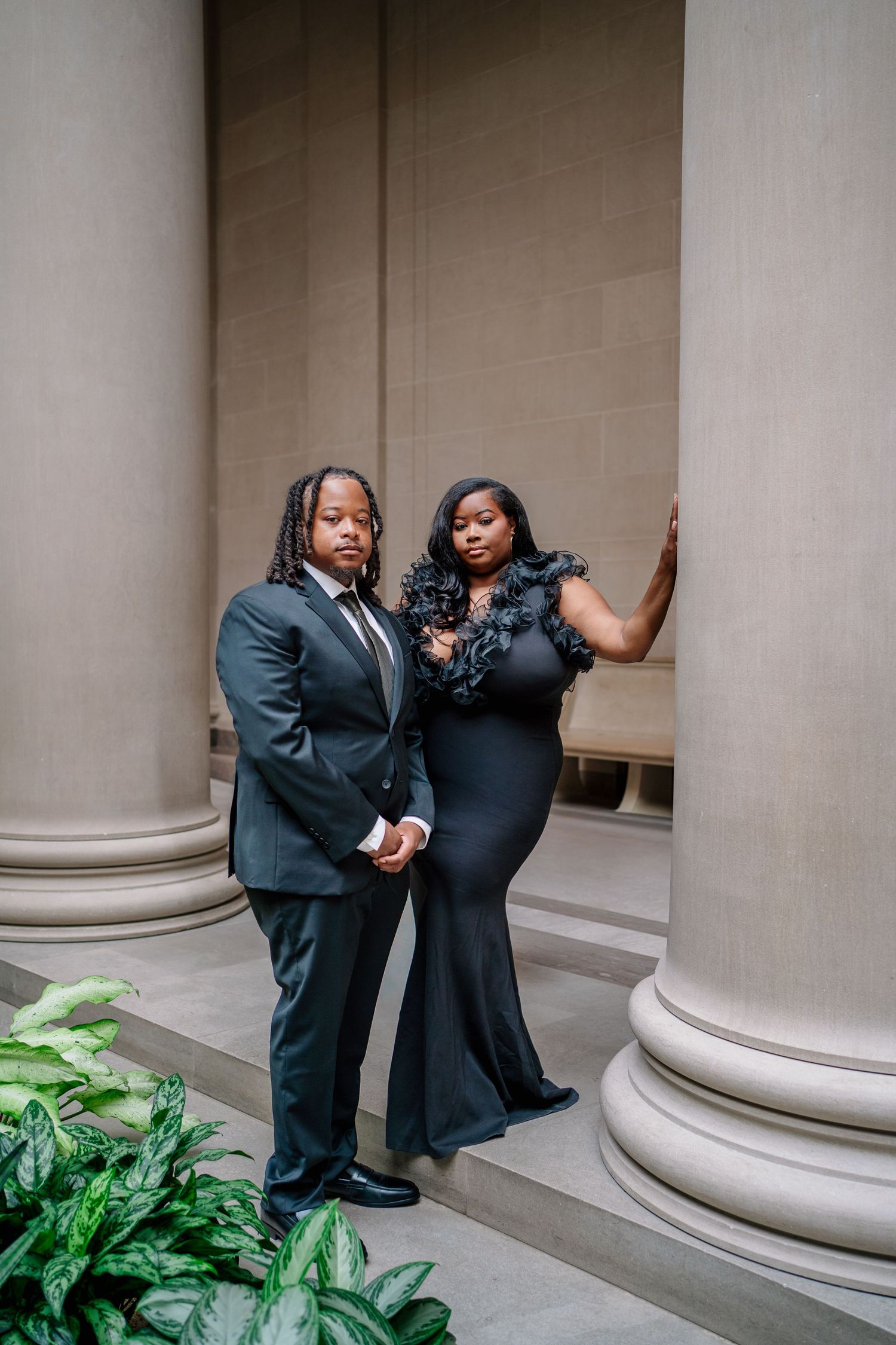Couple in elegant black attire posing between stone columns, woman in feathered gown and man in dark suit.