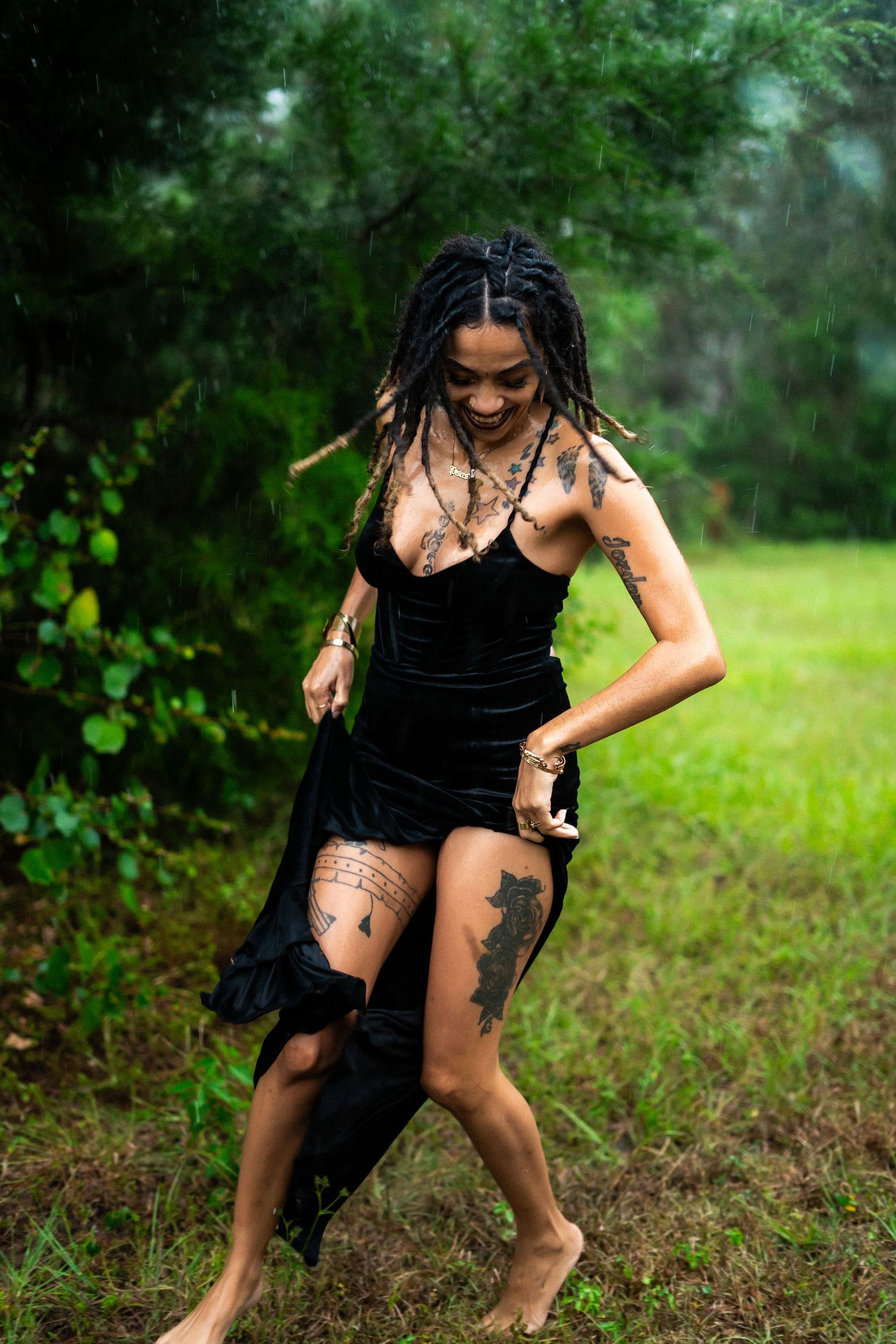 A person in a black dress poses outdoors in a grassy field with trees in the background.