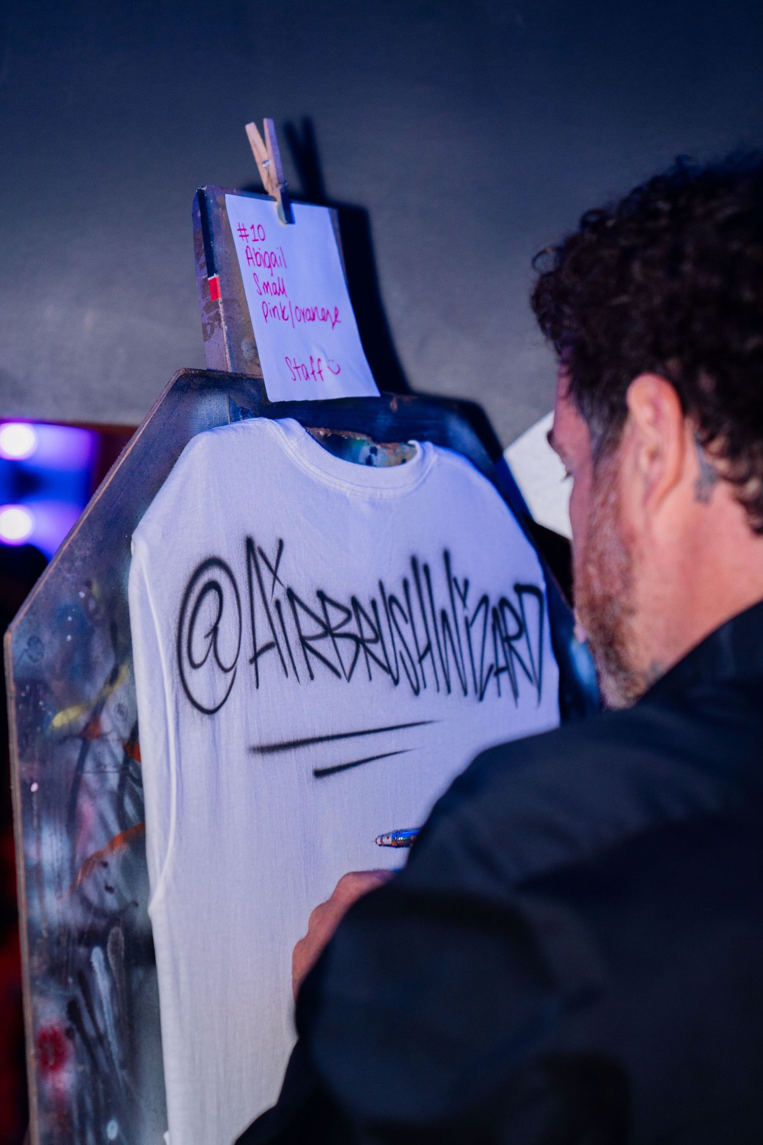 Artist airbrushing custom graffiti text on a white t-shirt at a creative event activation.