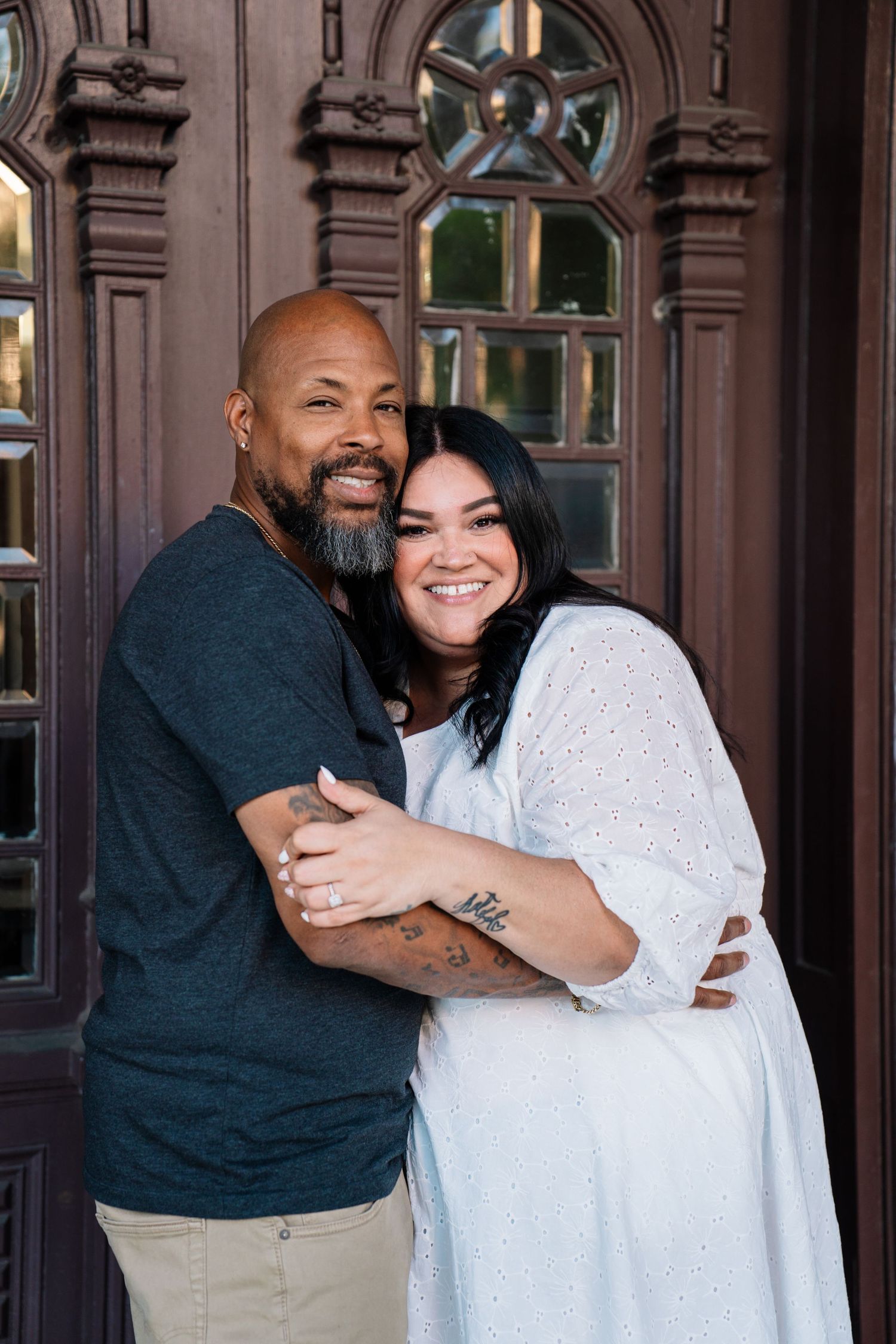 A smiling couple embraces in front of an ornate wooden door, the man bald with a beard, the woman in a white dress.